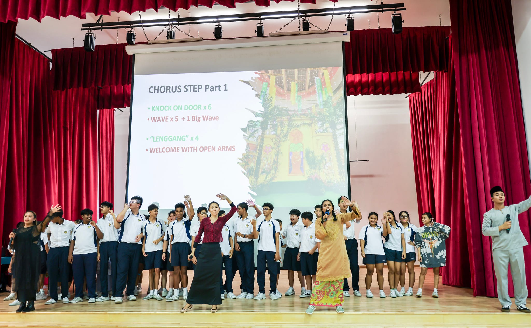 Students in uniform perform dance steps on a school stage with a projected slide showing "Chorus Step Part 1" instructions.