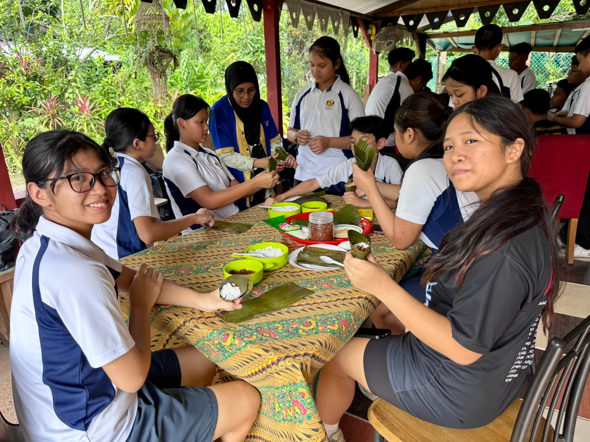 Students in school uniforms sit at a patterned table wrapping rice in banana leaves outdoors under a shelter.