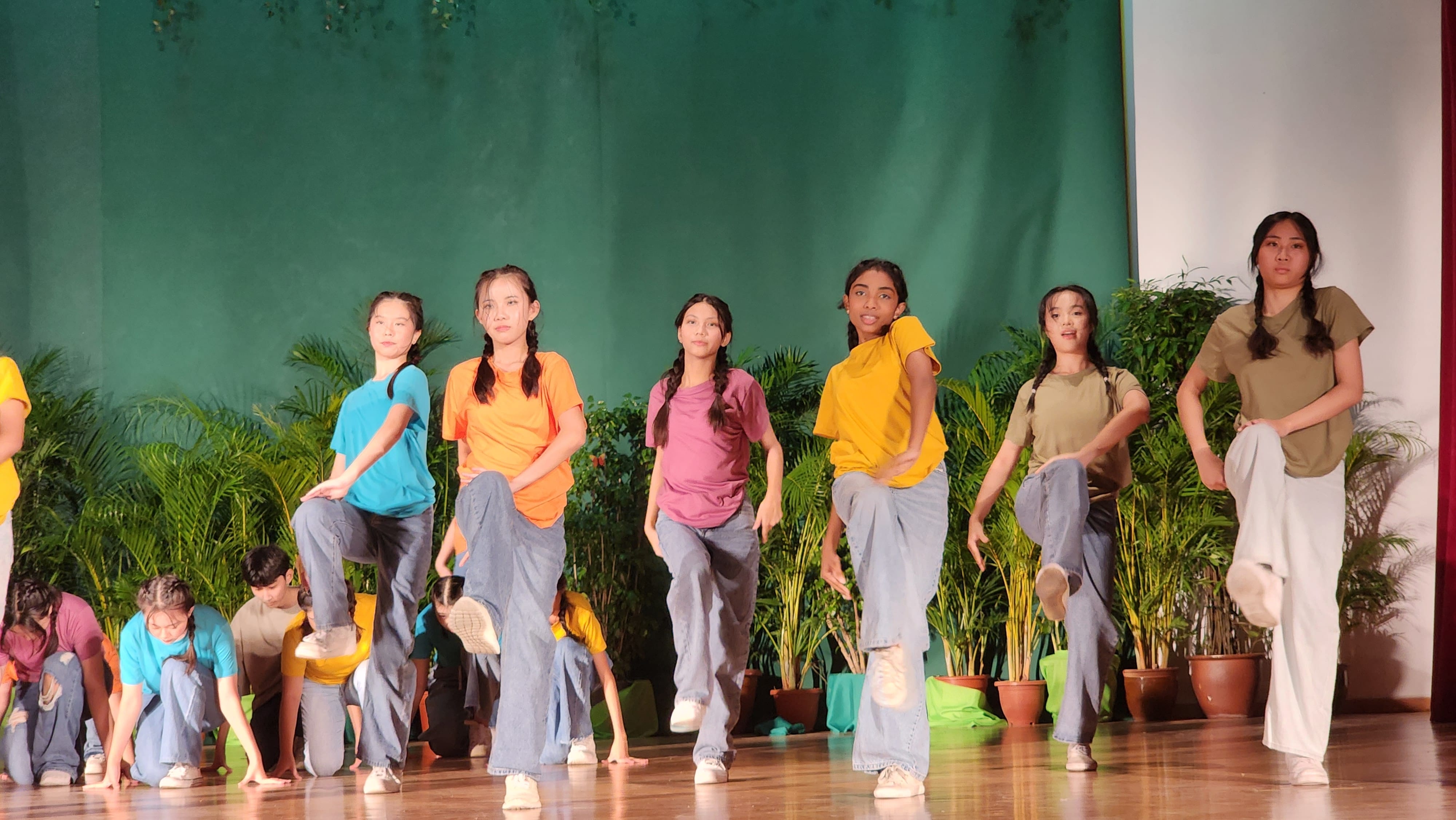 Girls in colorful t-shirts and jeans perform a synchronized dance on a wooden stage with green backdrop and plants.