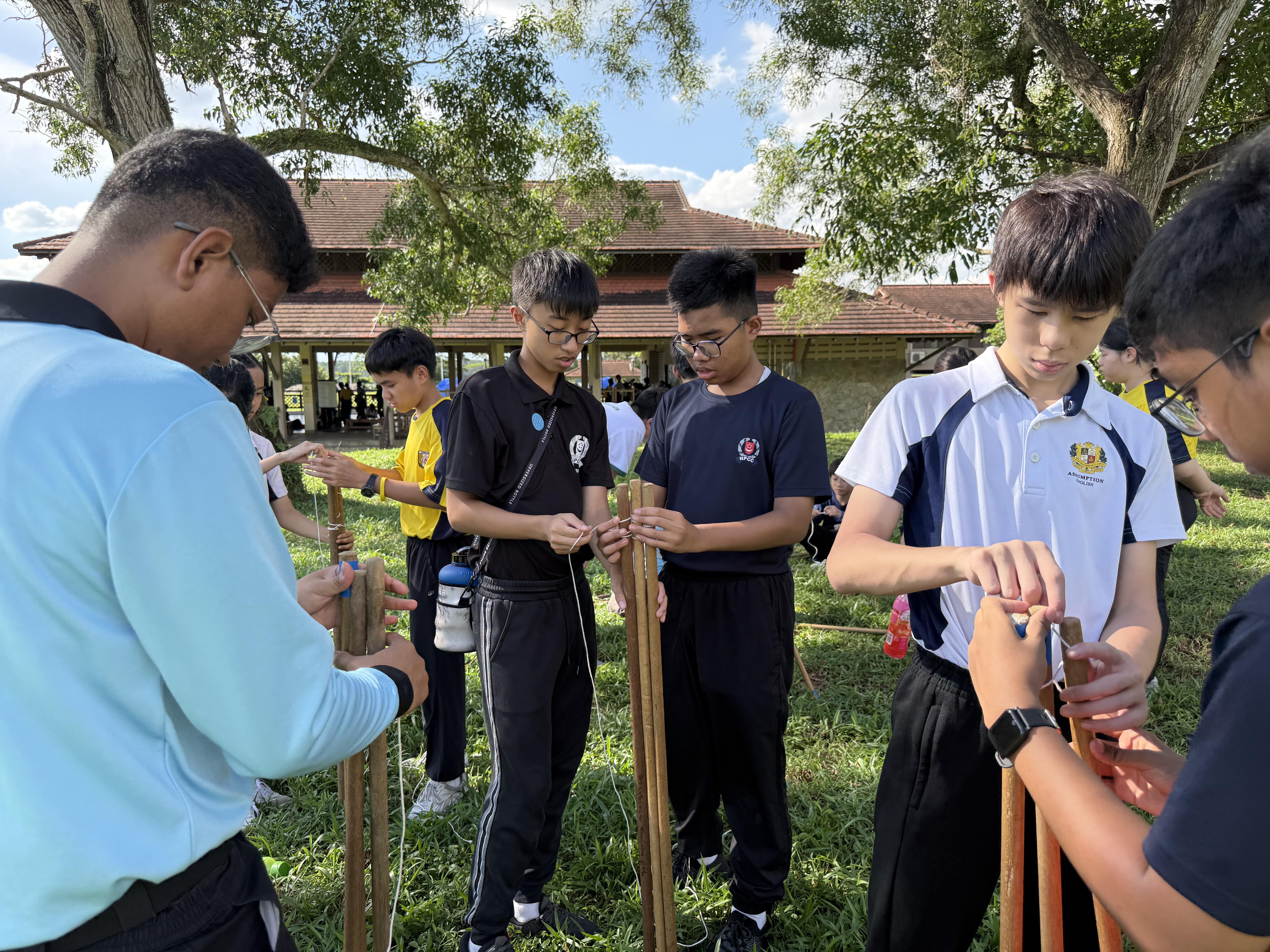 Several students in school uniforms stand outdoors tying string around wooden poles on a grassy field.