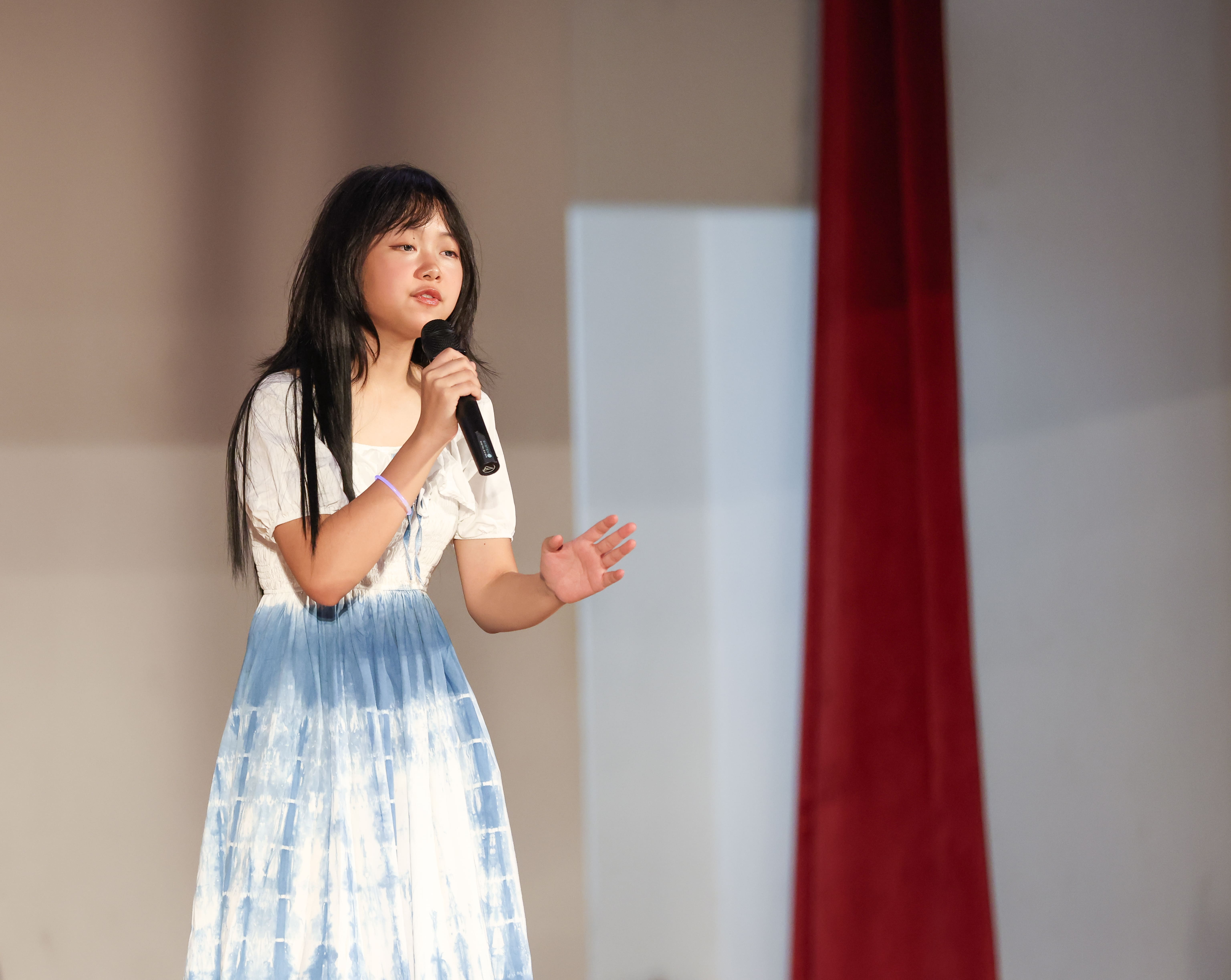 Young girl with long black hair in a blue tie-dye dress holds a microphone and gestures on stage.