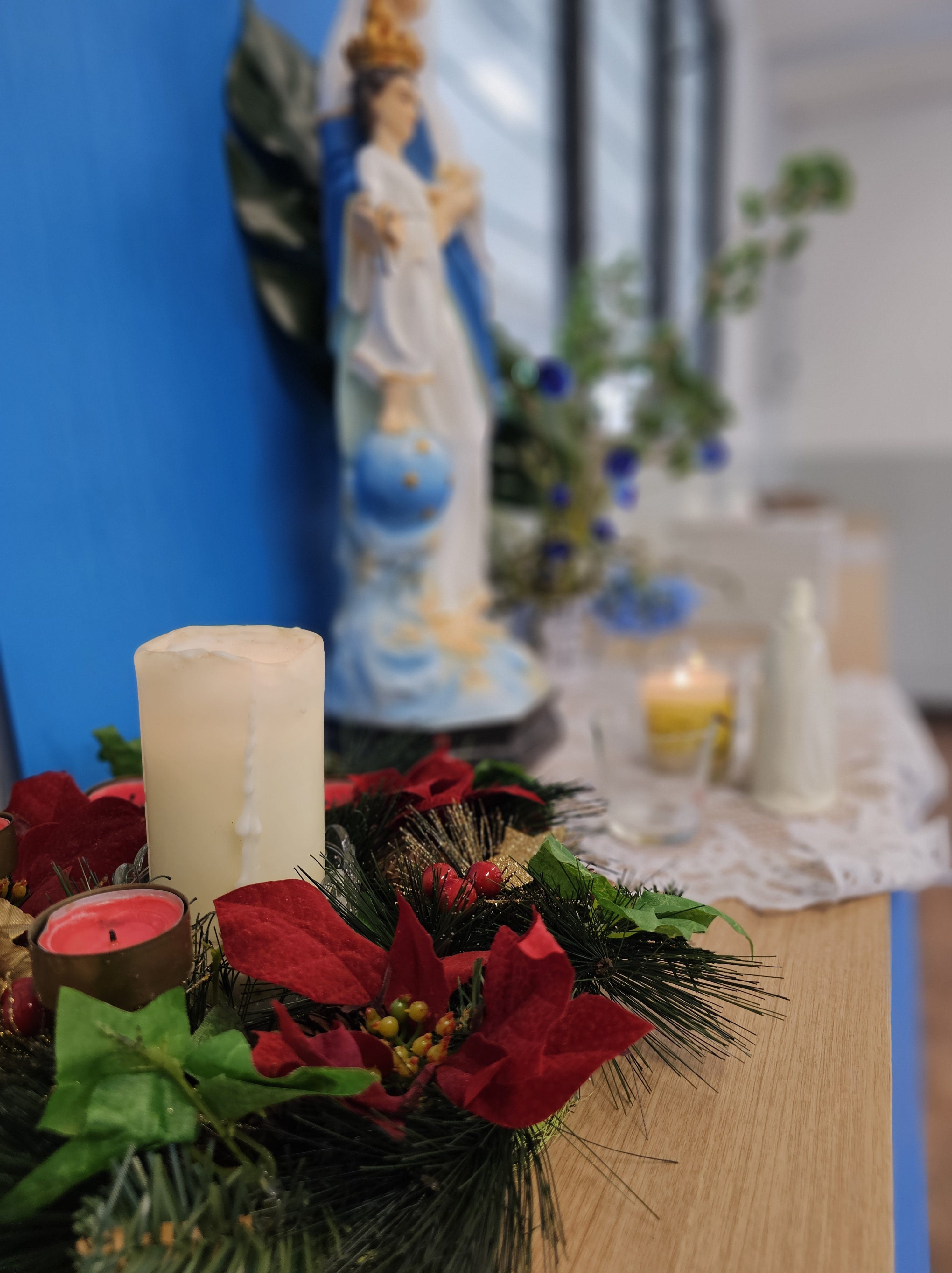 A white pillar candle in a red poinsettia wreath on a table, with a blurred Virgin Mary statue behind it.