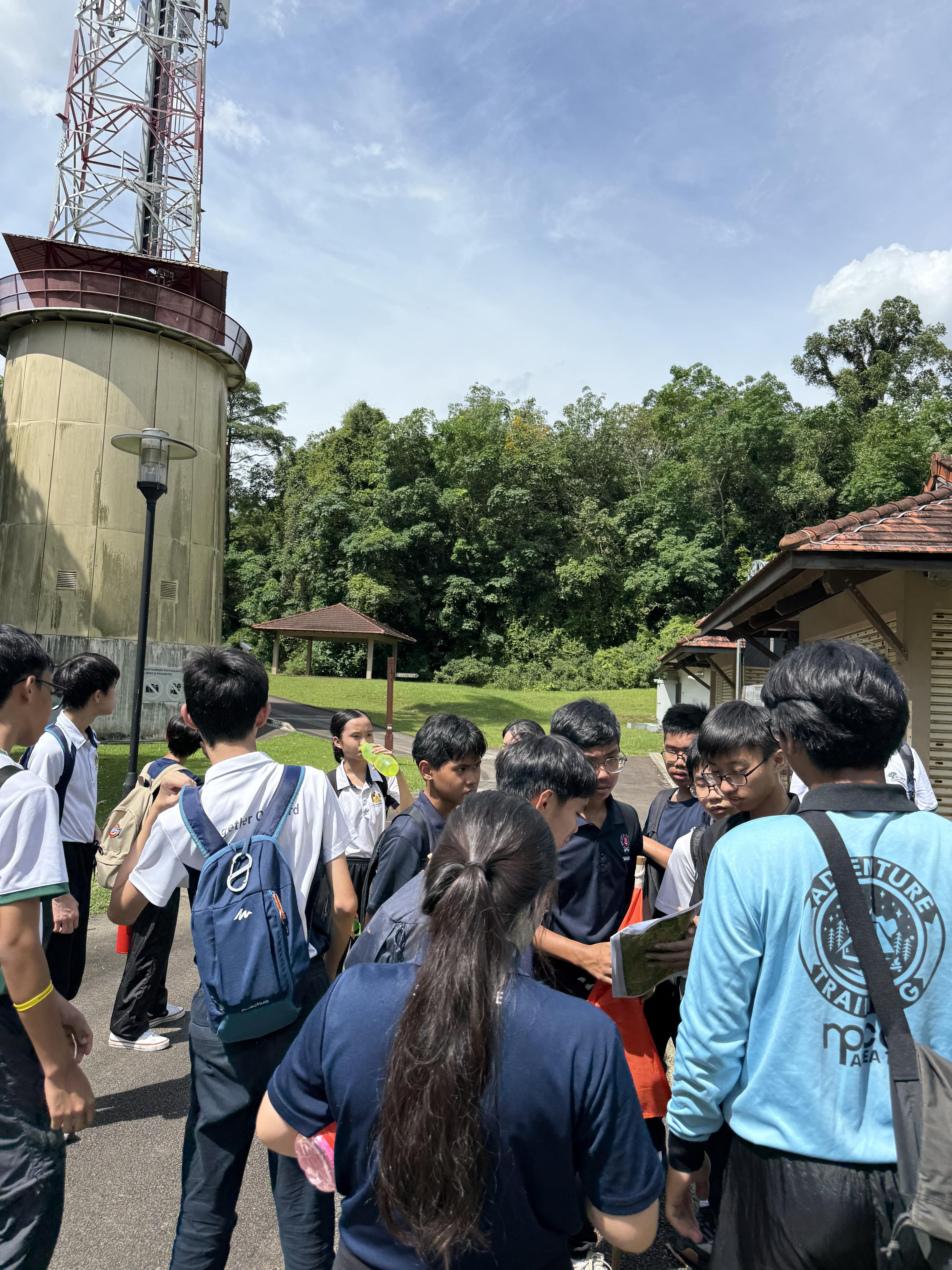A group of students in uniforms gather outdoors near a tall tower and small buildings, surrounded by trees.