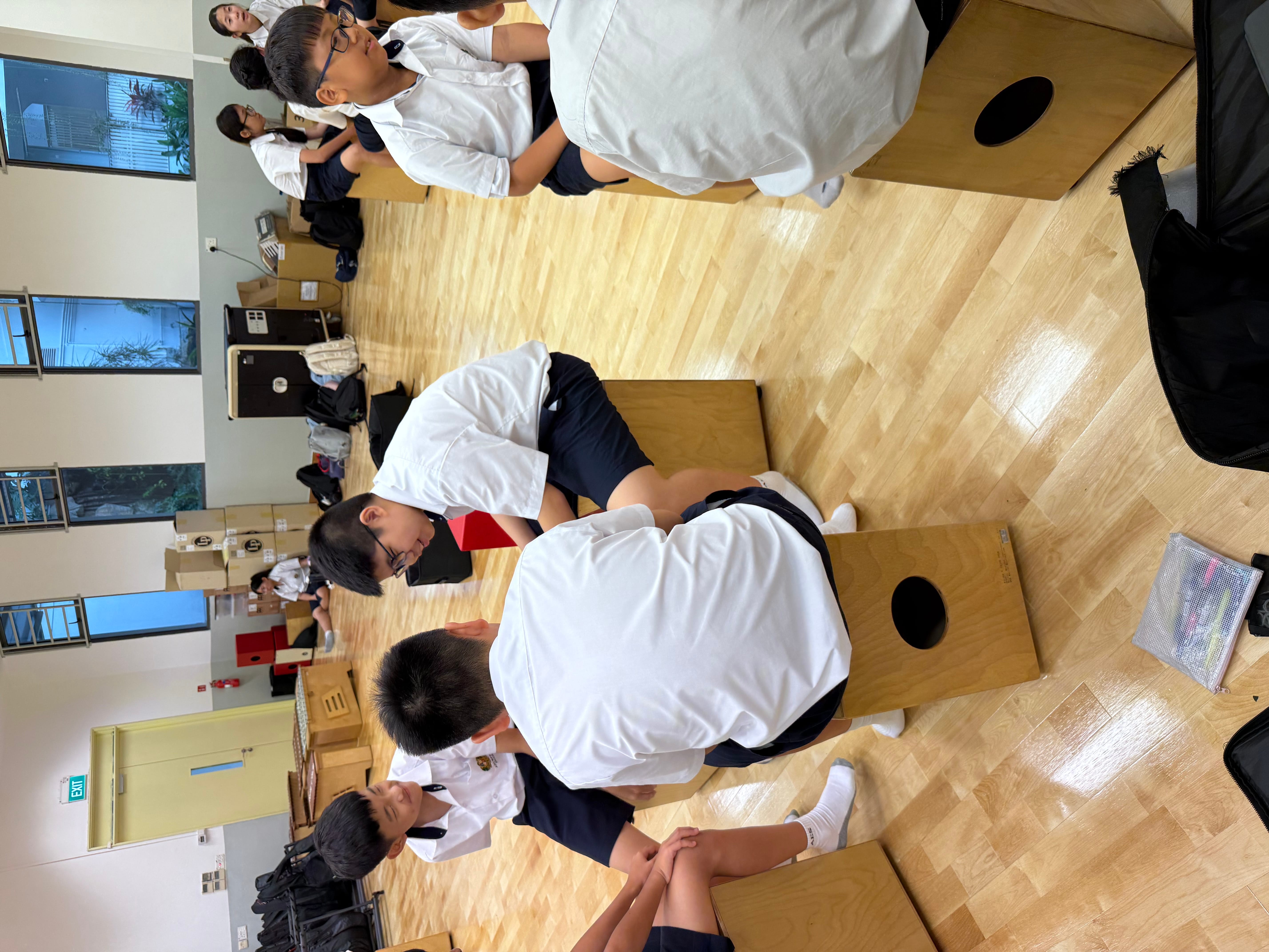 Students in white uniforms sit on wooden cajon drums in a music room with a polished floor.