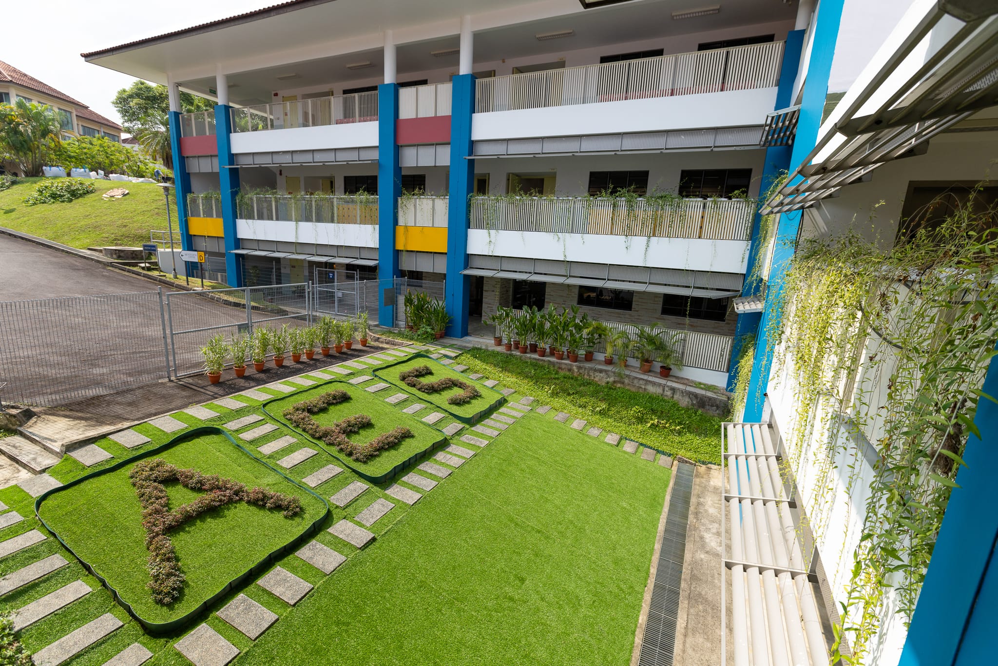 A multi-story school building with blue columns, balconies, and a courtyard featuring letters spelled out in shrubs on grass.