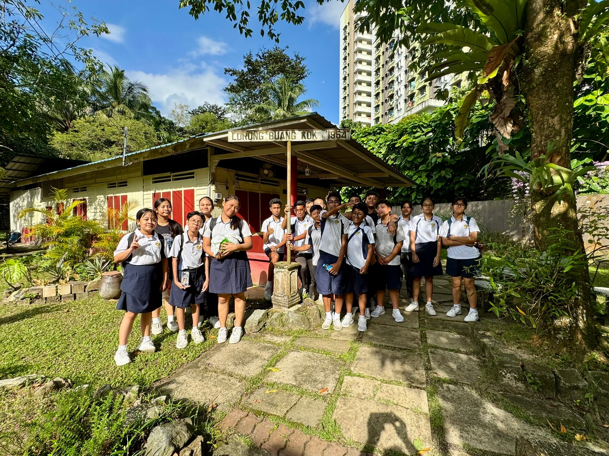 A group of students in white and navy uniforms stand outside an old building with a sign reading Lorong BuangKok 1964.