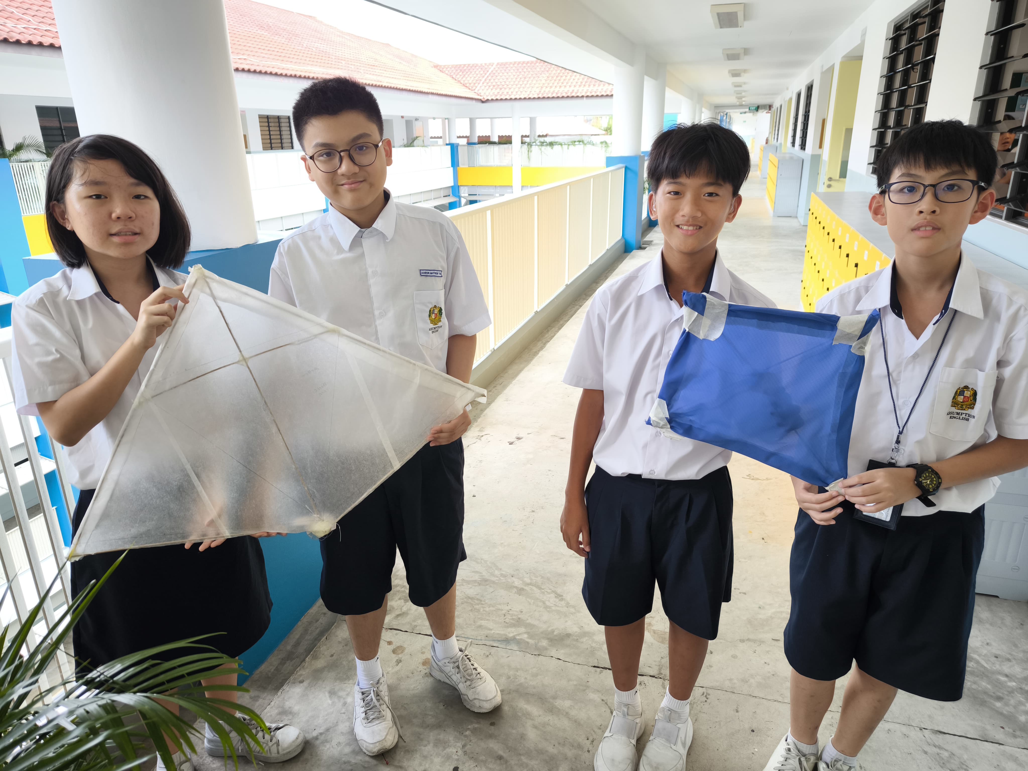 Four students in white school uniforms hold handmade kites, one clear and one blue, in a school corridor.