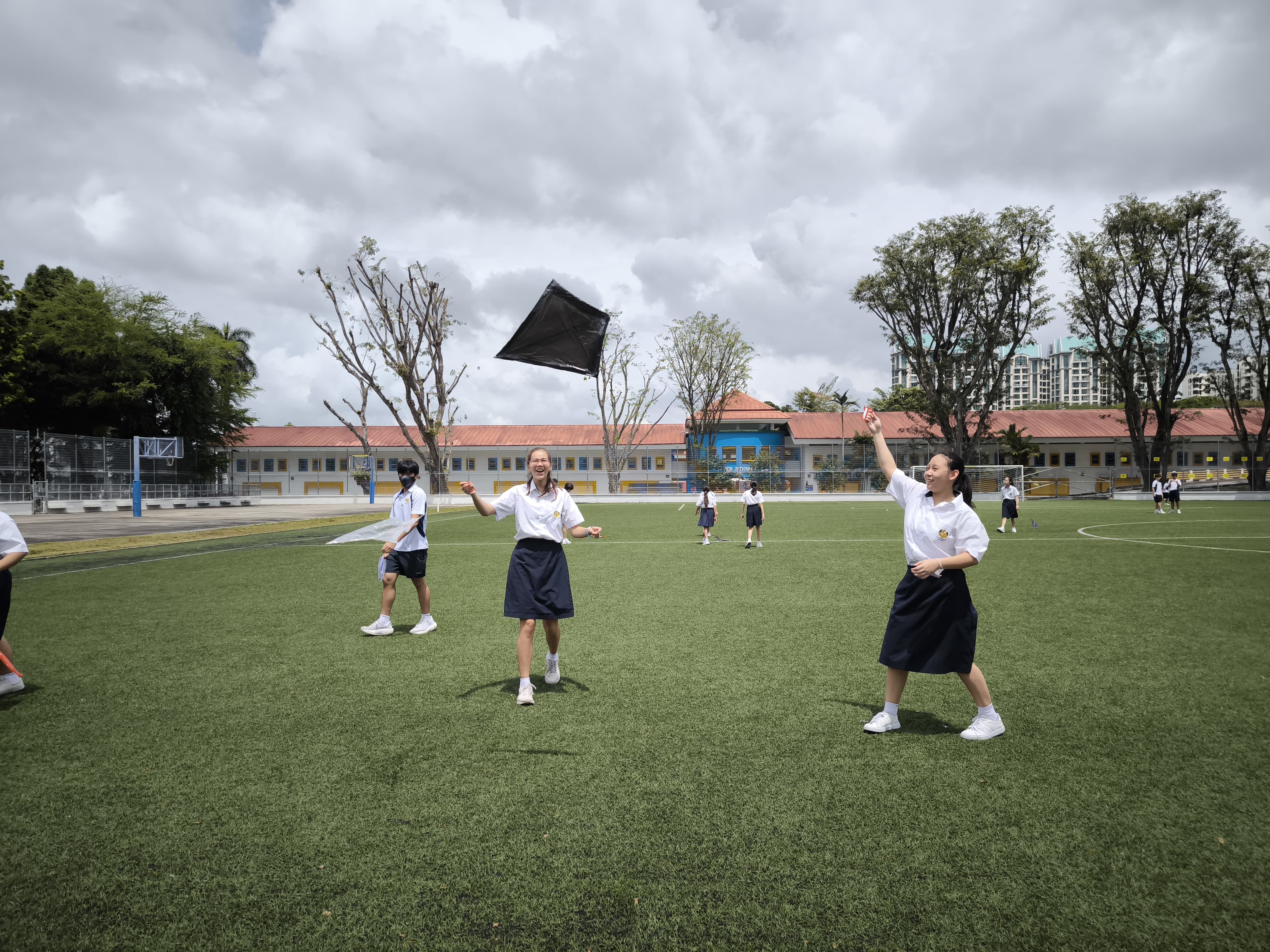 Students in white shirts and dark skirts flying a black kite on a green school field under a cloudy sky.