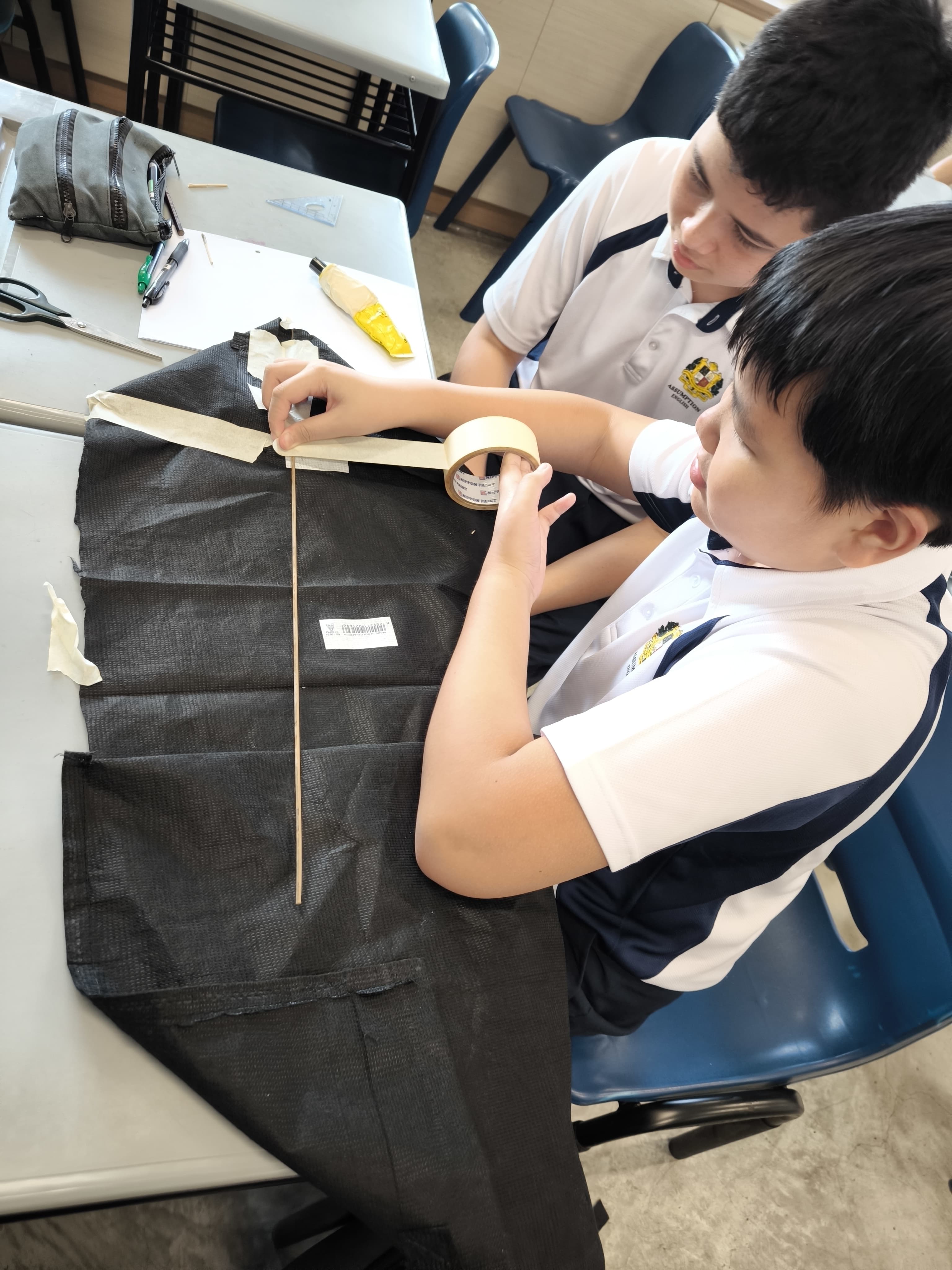 Two school boys in uniforms applying masking tape to a wooden stick on black fabric at a classroom desk.