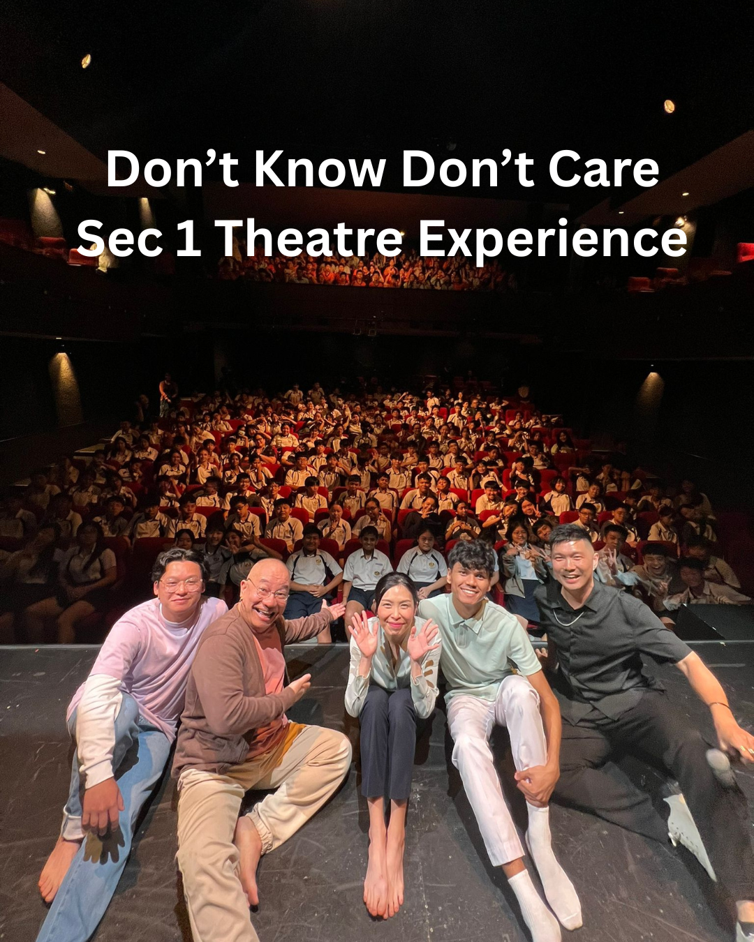 Five performers sit on a stage in front of a packed theatre audience of uniformed students in red seats.