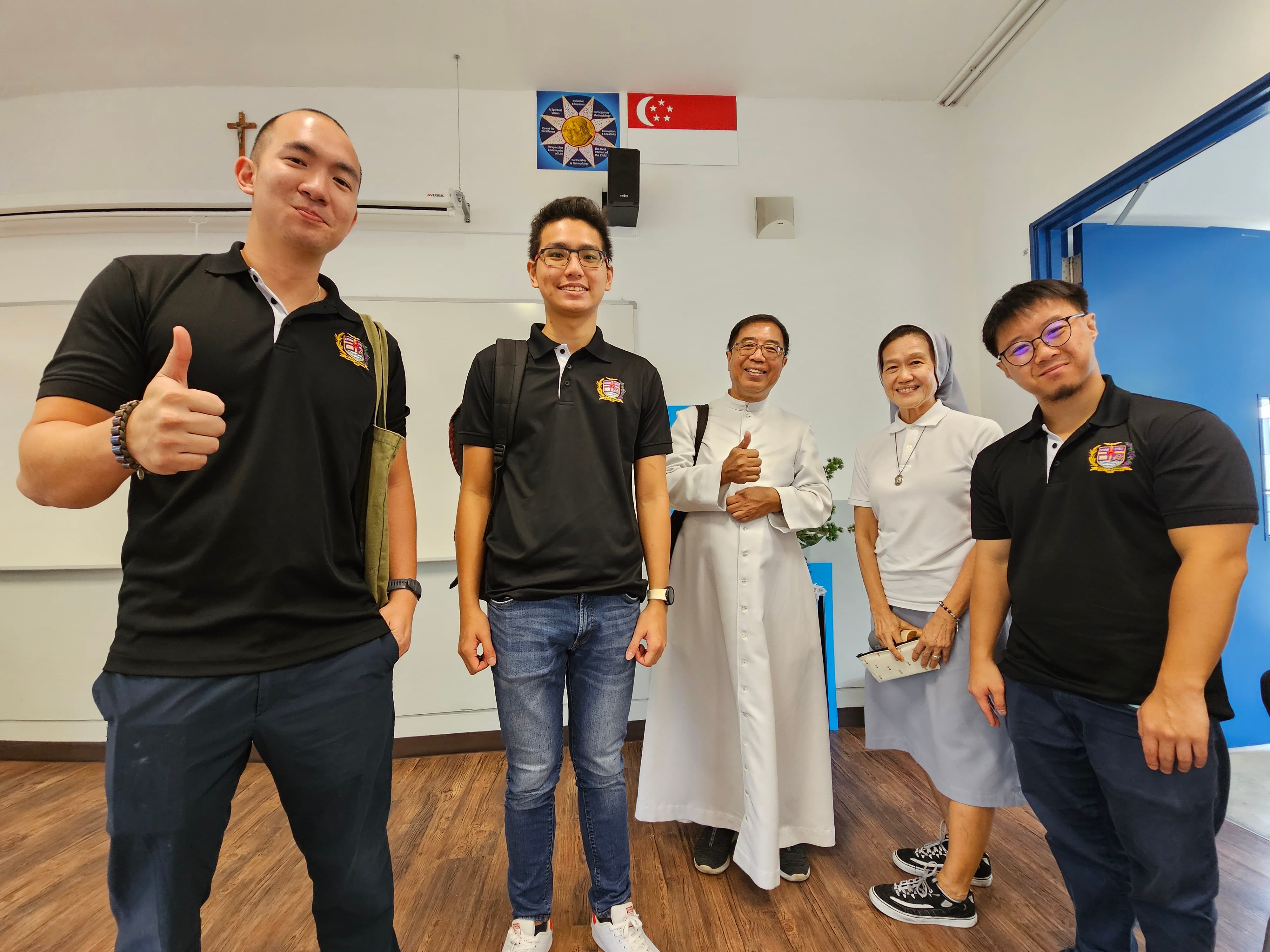 Five people pose smiling in a classroom with a Singapore flag and cross on the wall; two wear white religious habits.