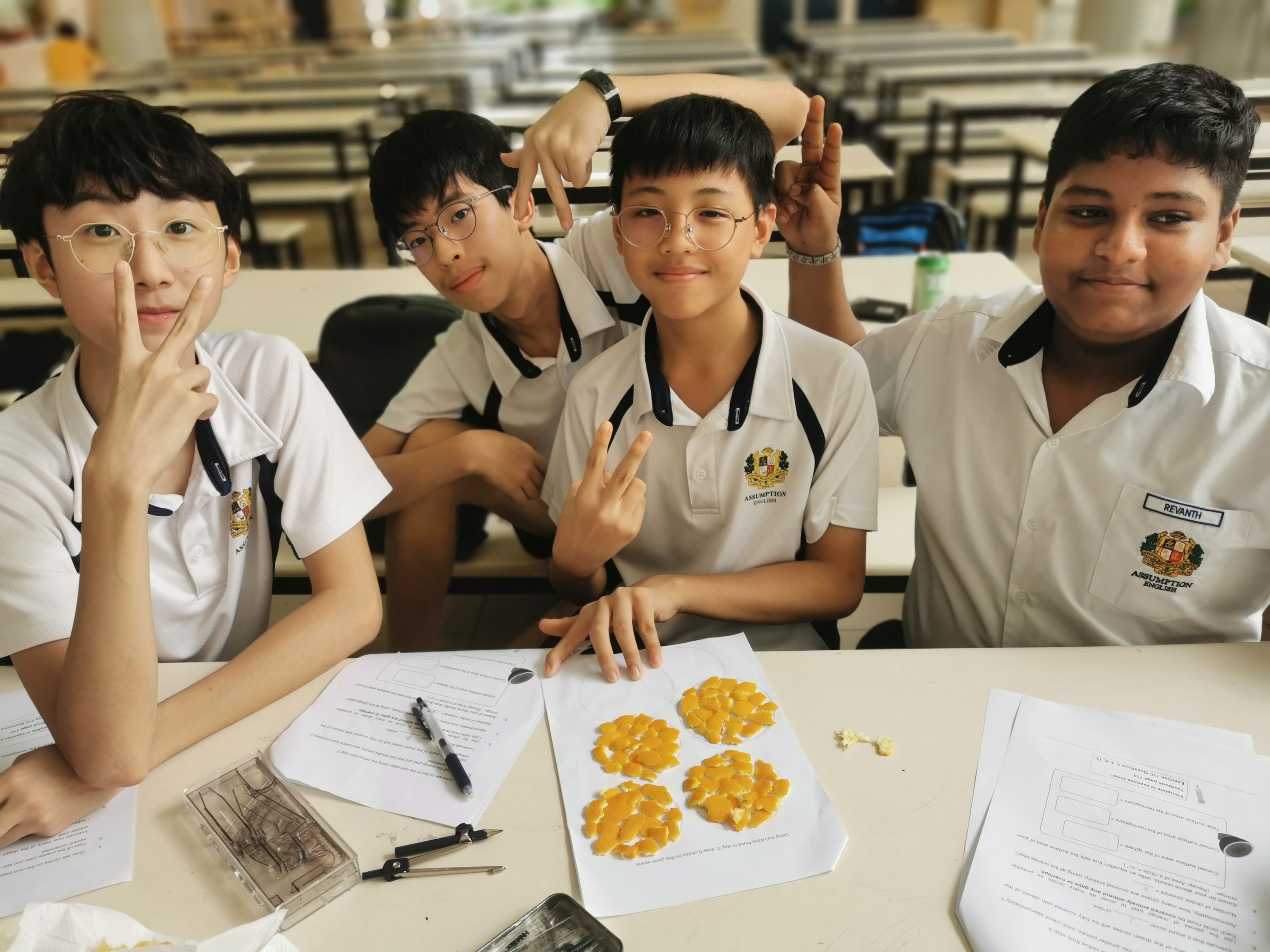 Four school students in white uniforms pose playfully at a desk with orange fruit segments arranged on paper.