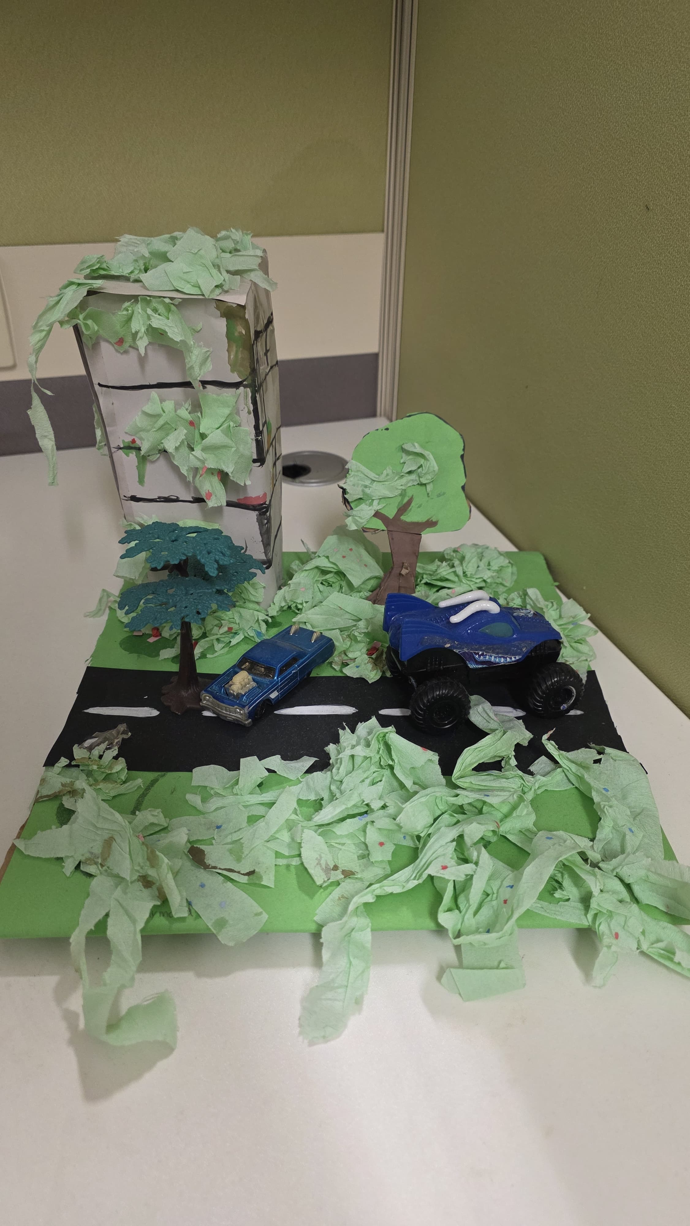 A handmade diorama with toy cars on a paper road, green tissue paper grass, cardboard trees, and a building.