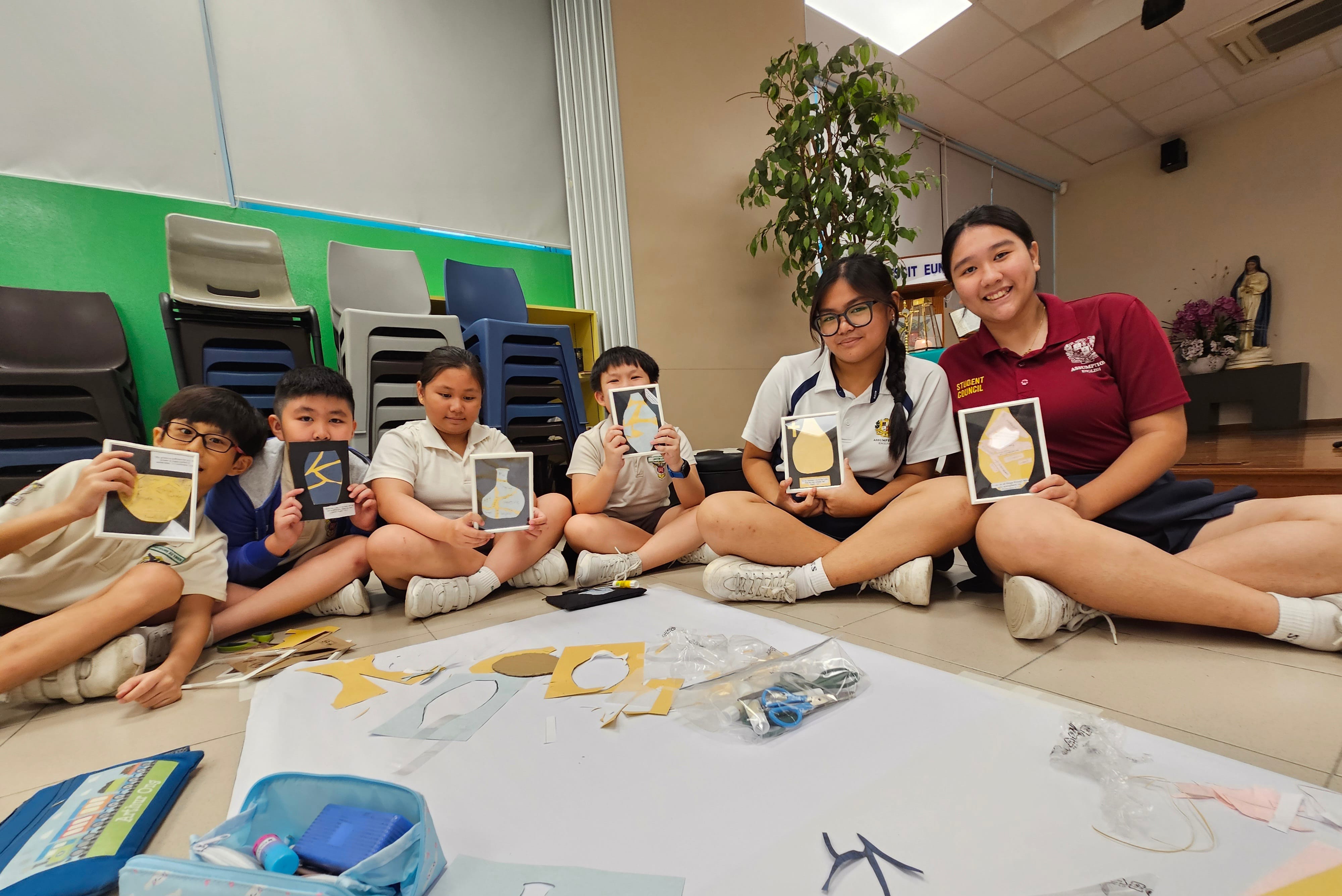 Six students sit on a floor holding up small art cards with gold designs, surrounded by craft materials.