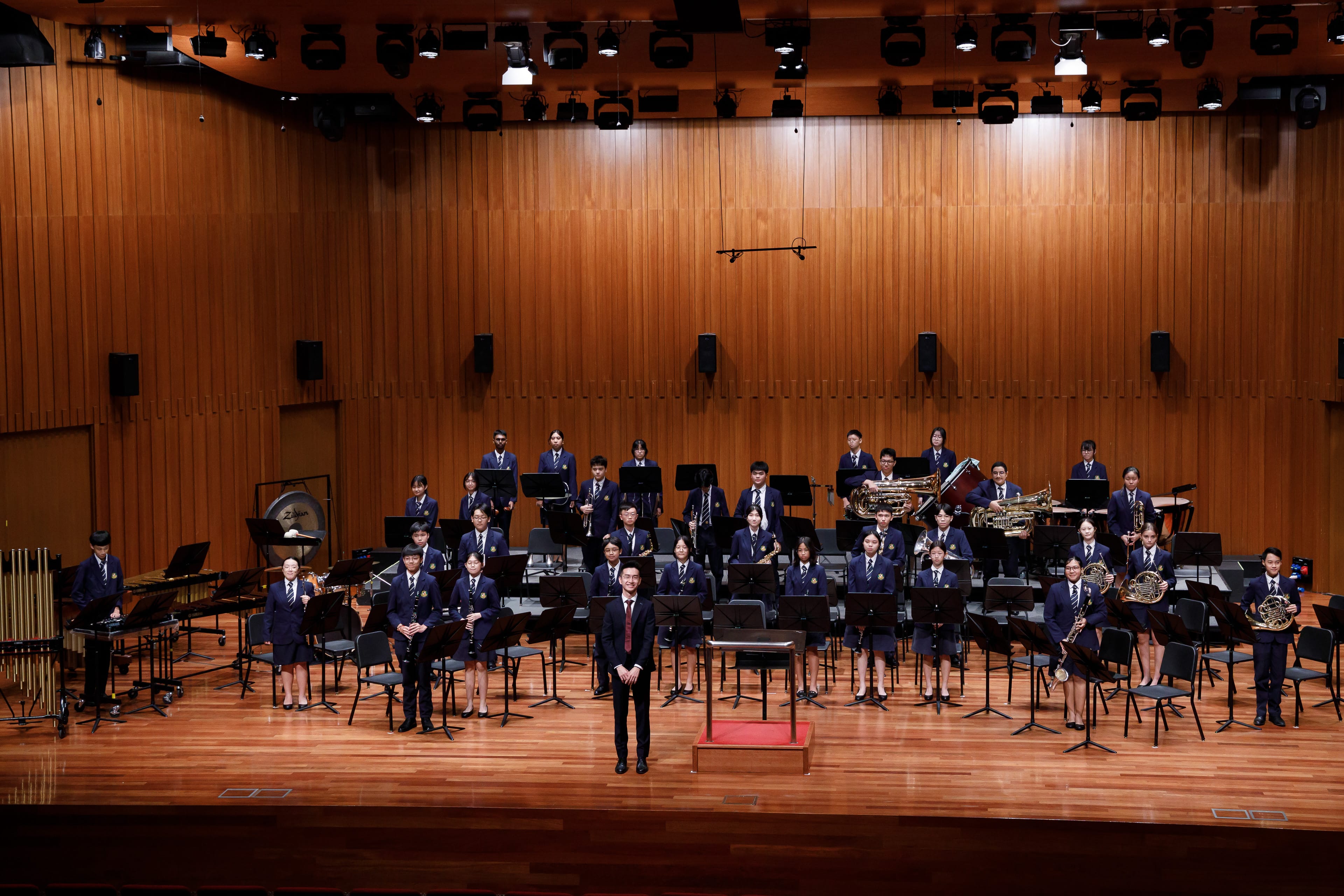 A youth wind band in navy uniforms poses on a wooden concert stage with a conductor standing in front.