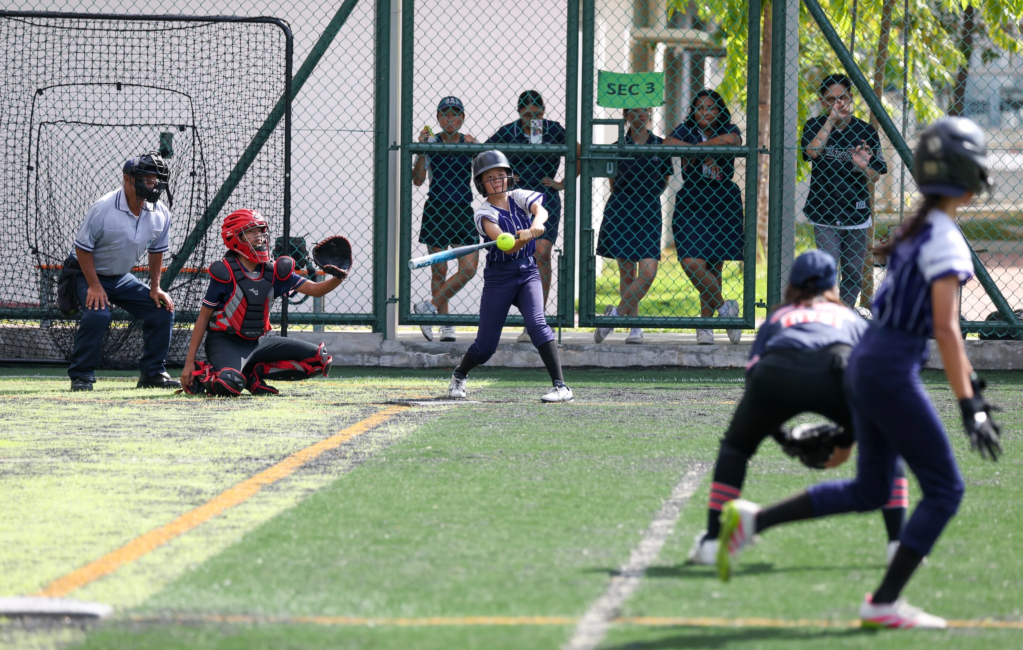 A batter in purple swings at a softball pitch as catcher, umpire, and spectators watch behind a chain-link fence.