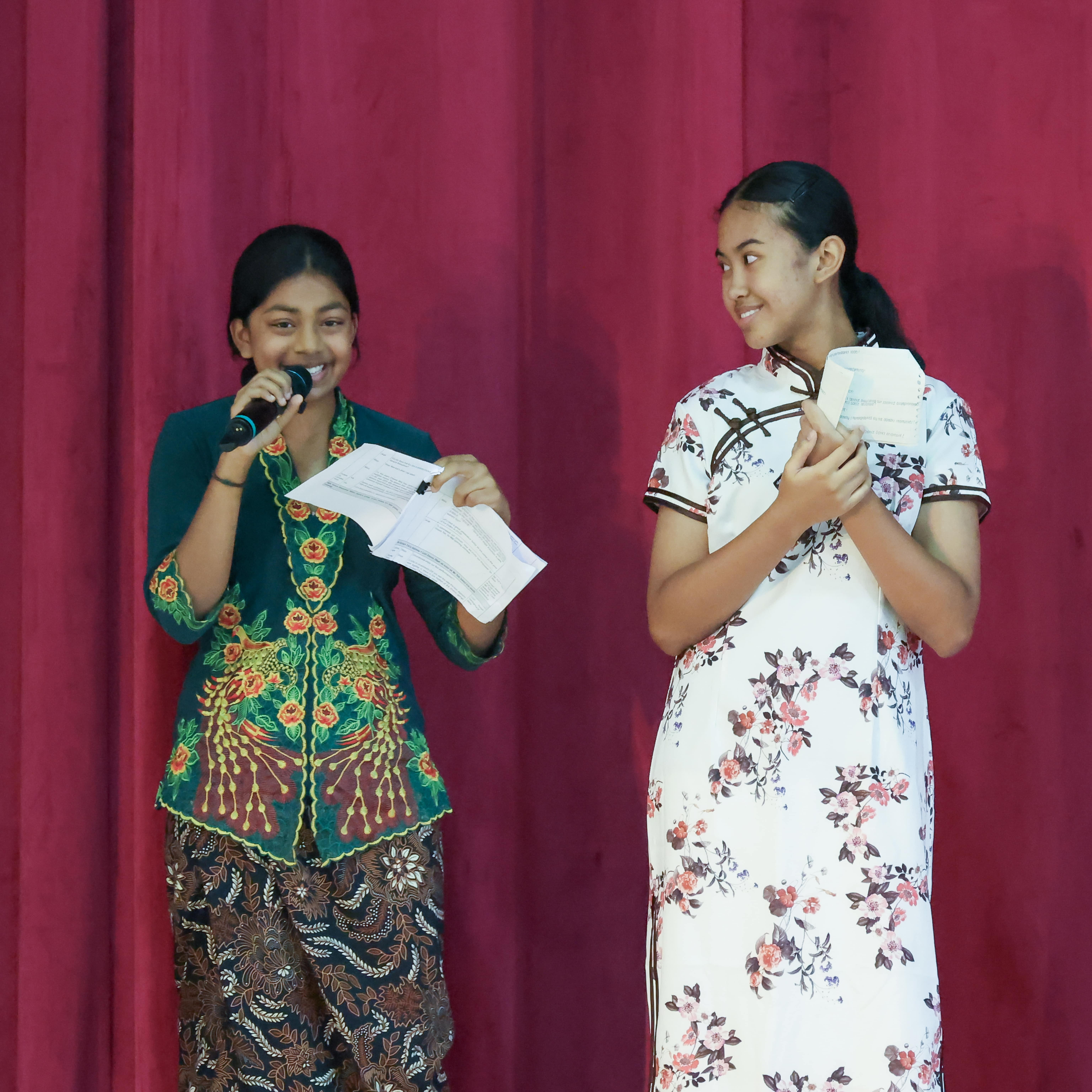 Two girls in traditional cultural outfits stand on stage, one speaking into a microphone holding papers, before a red curtain.