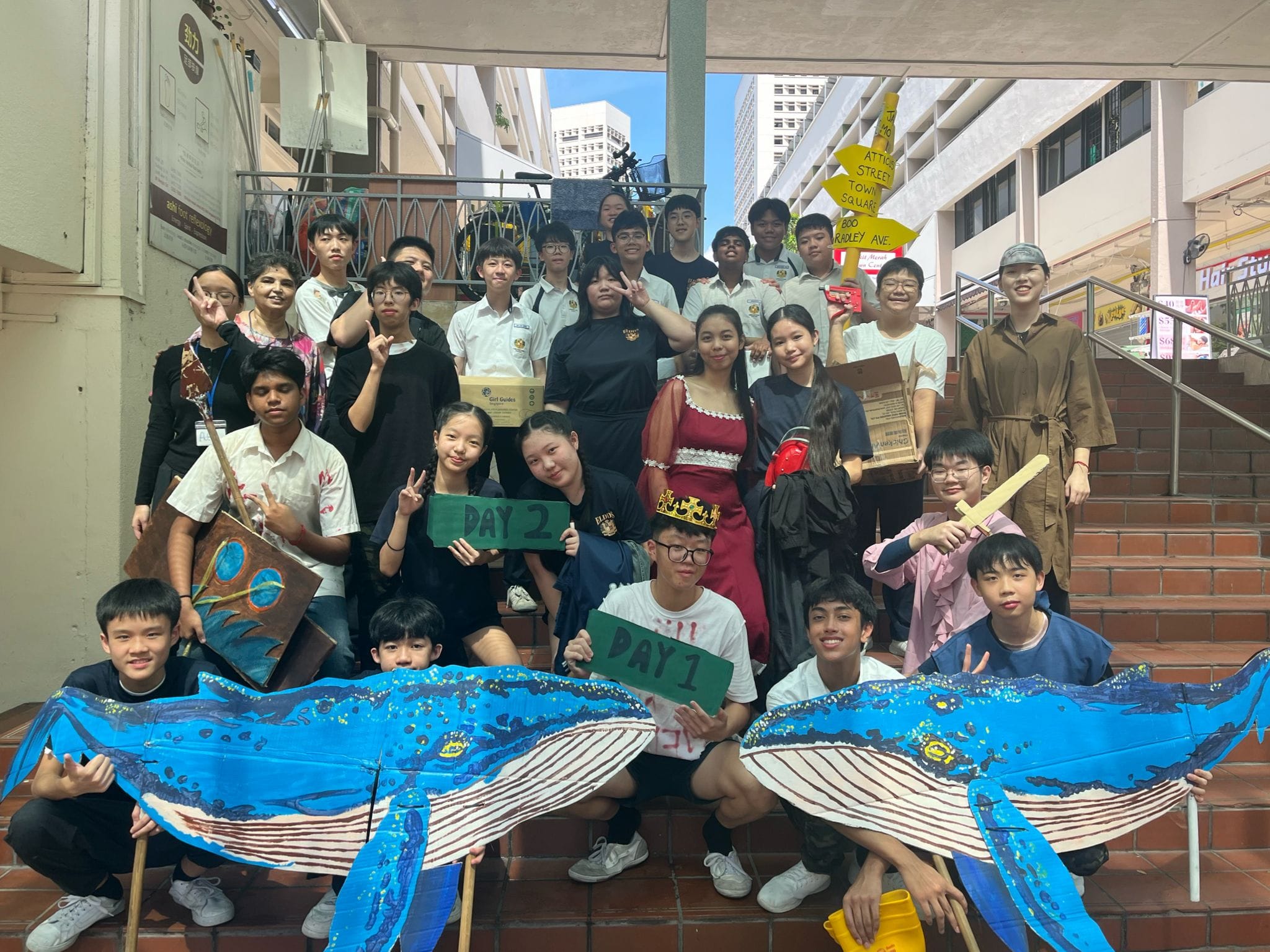 A large group of students pose on outdoor steps holding a painted whale banner and signs reading Day 1 and Day 2.
