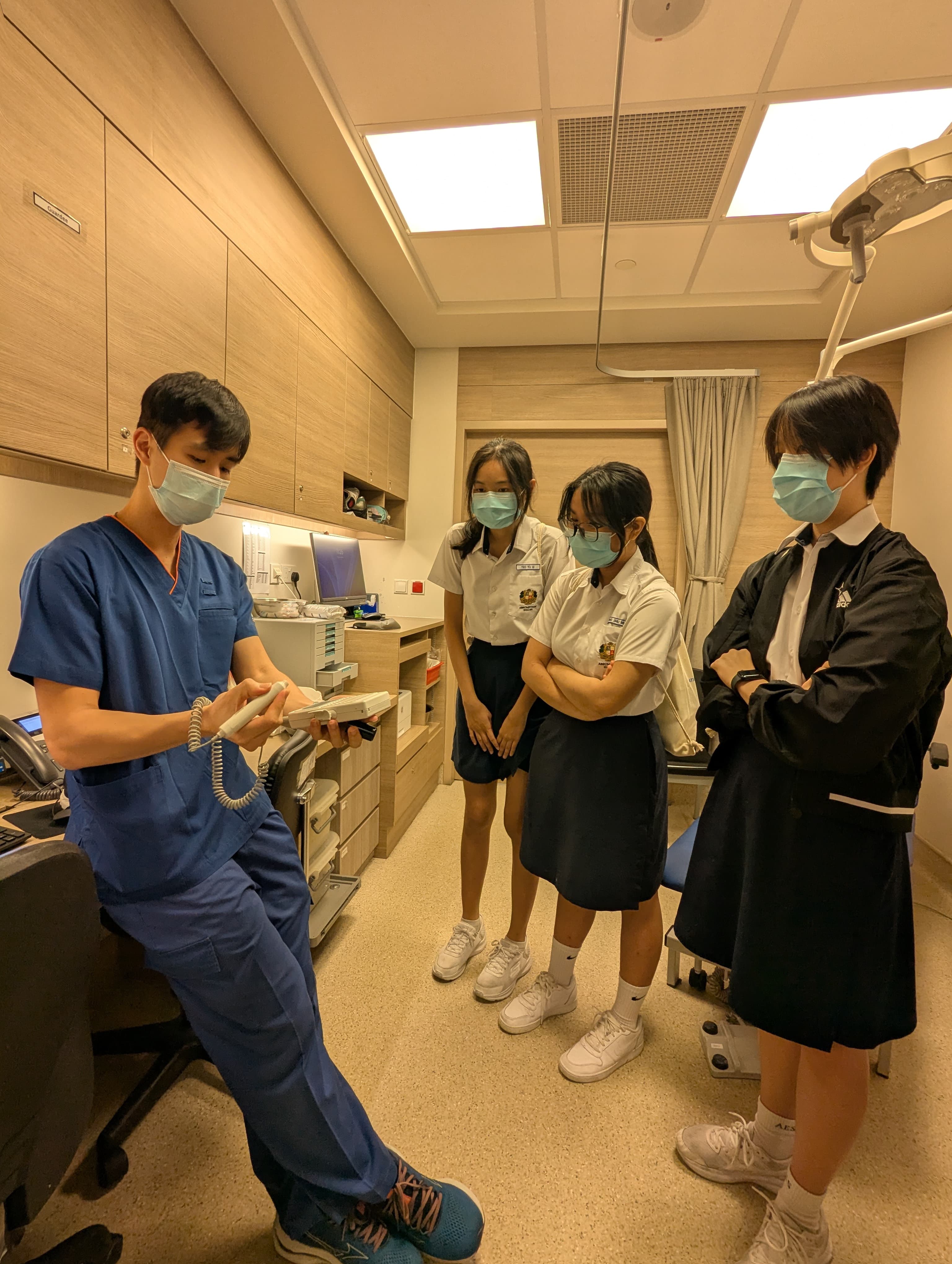 A masked dental professional in blue scrubs shows a tool to three masked students in school uniforms inside a clinic room.