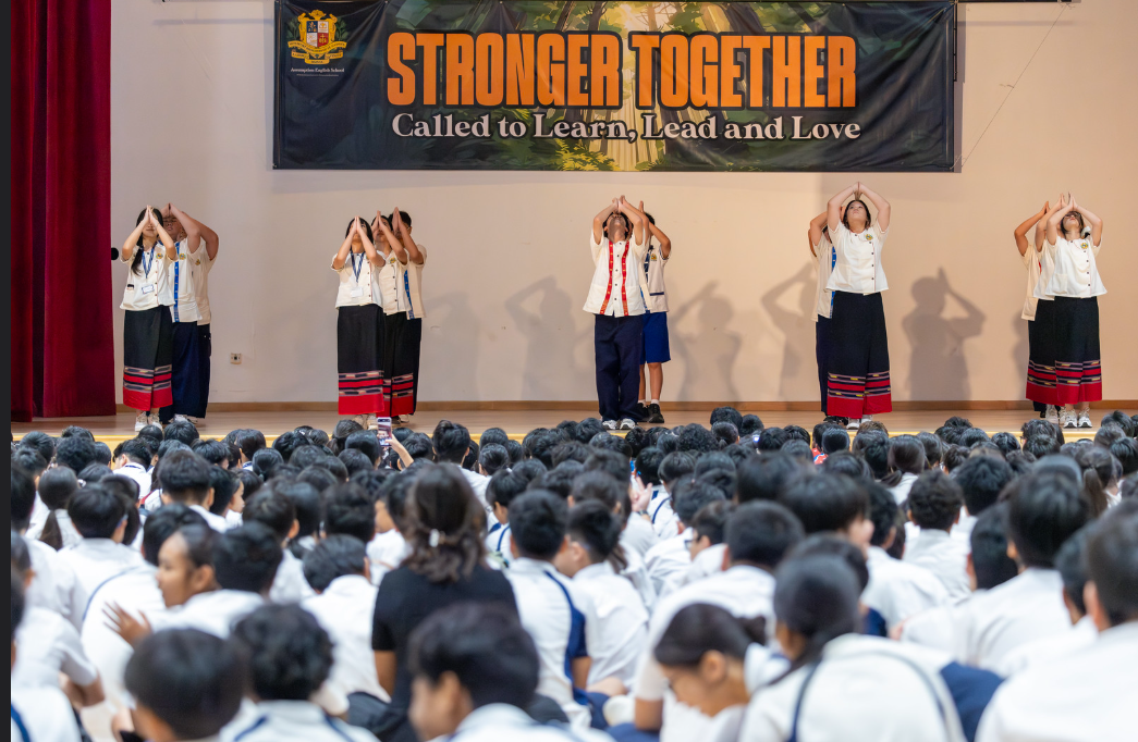 Students perform on stage with arms raised before a seated audience under a "Stronger Together" banner.