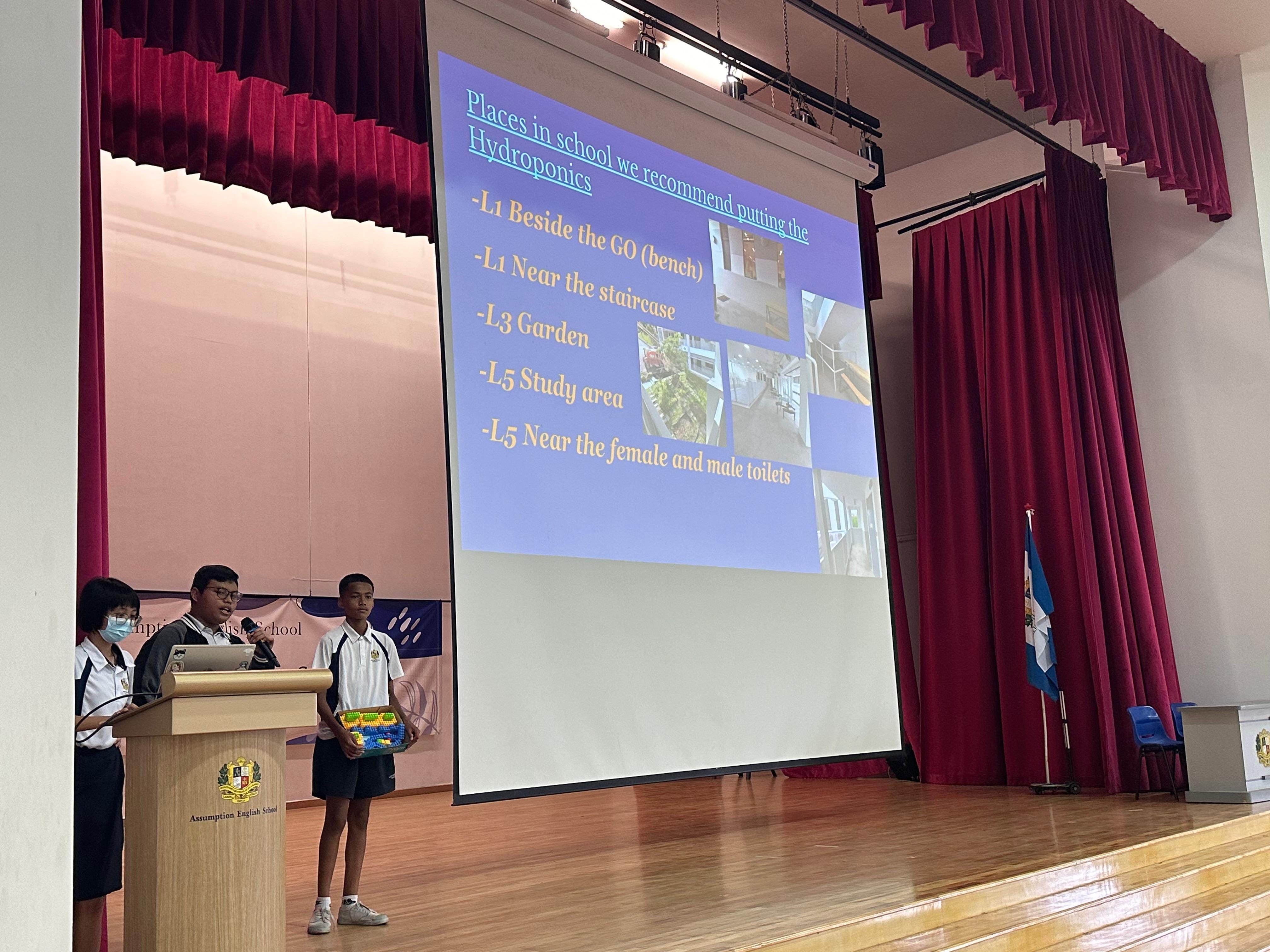 Three students present on stage at Assumption English School, with a slide about hydroponics locations displayed behind them.