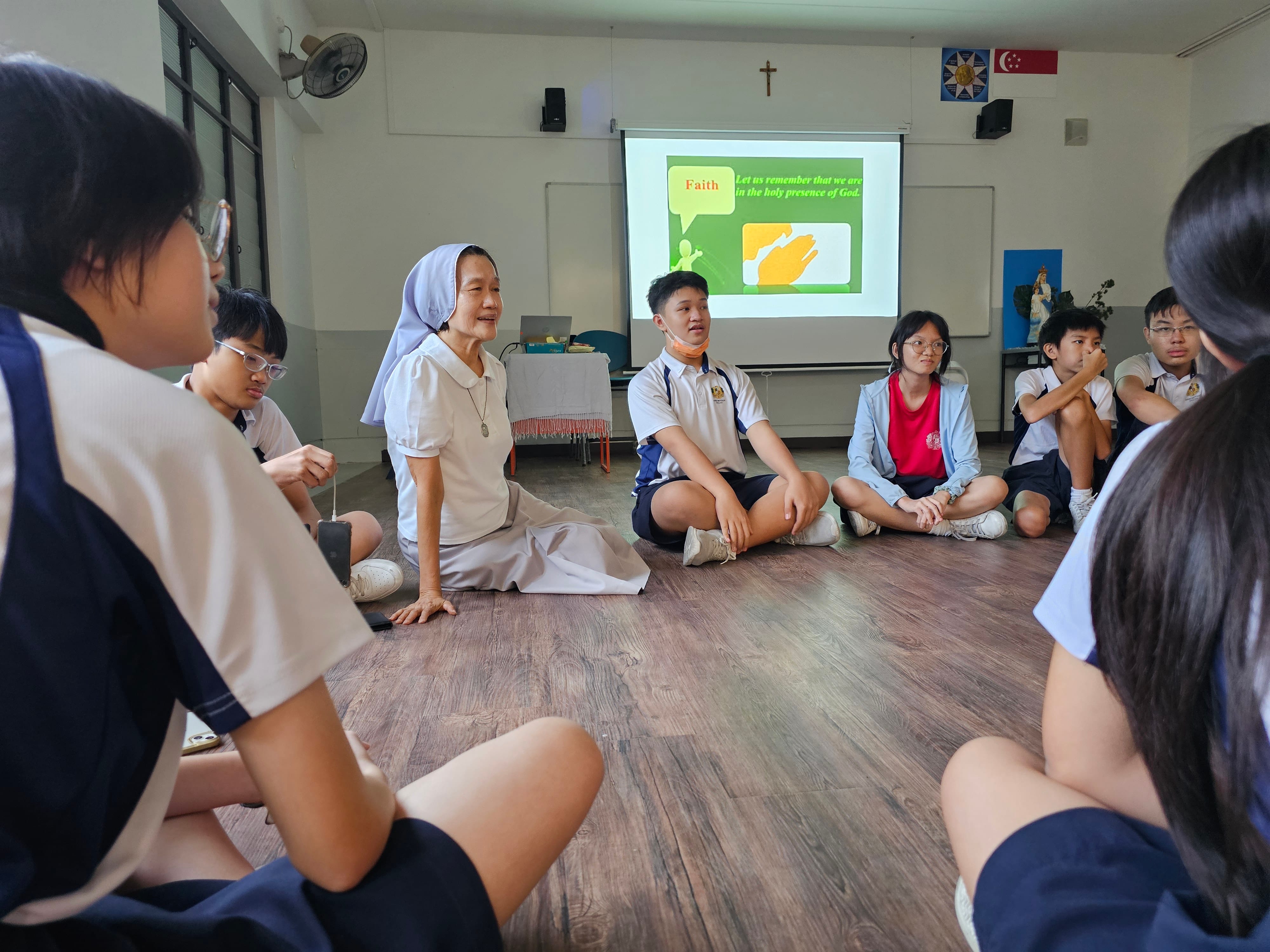 A nun in white habit sits on the floor with school students in a circle during a Faith lesson shown on a projector screen.
