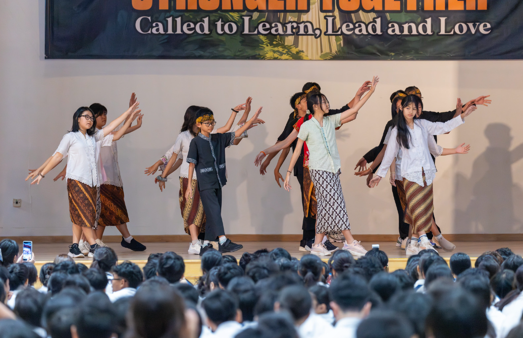 Students in traditional batik skirts perform a dance on stage before a large seated audience under a school banner.