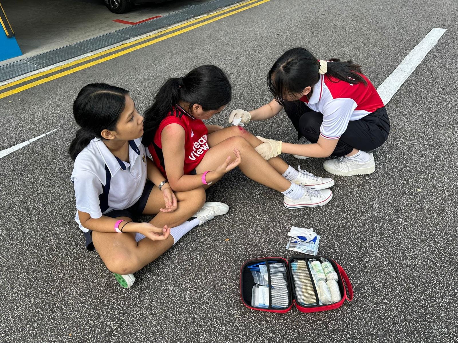 Three girls on pavement as one tends to another's injured knee using gloves, with an open first aid kit nearby.