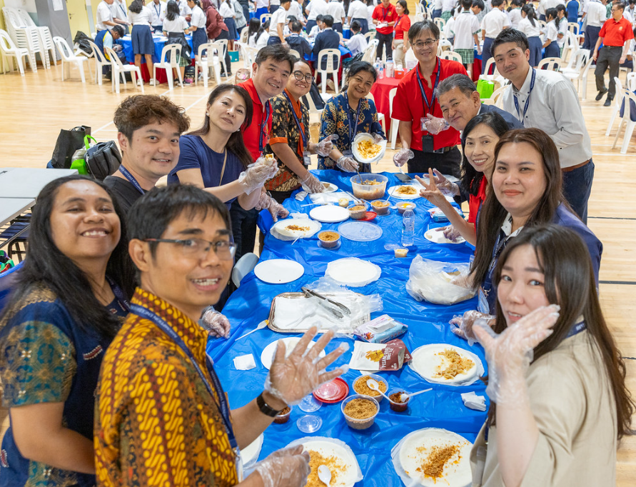 A group of smiling adults wearing plastic gloves sit around a blue-covered table with food in a large hall.