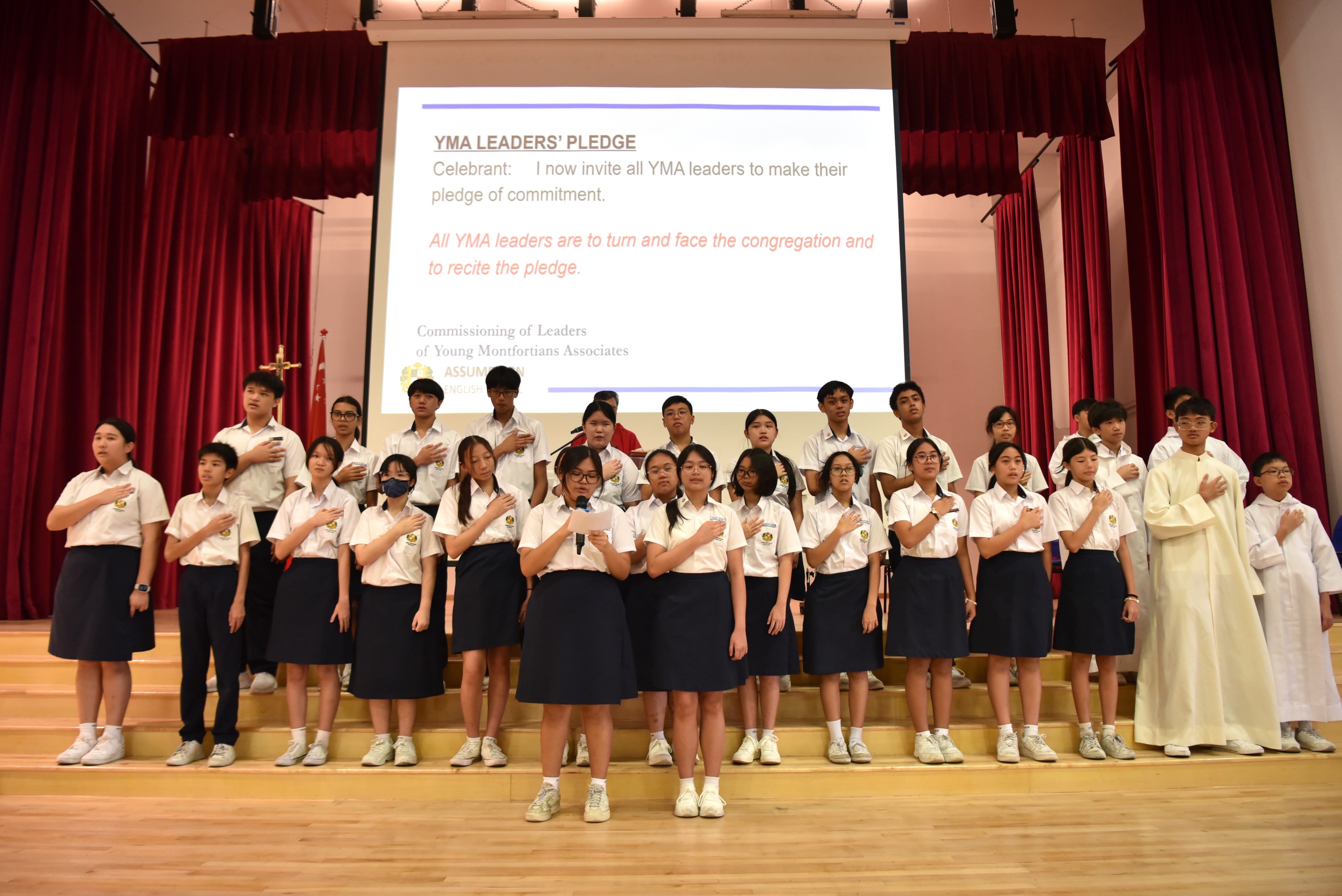 Students in white uniforms stand on a stage with hands on chests reciting the YMA Leaders' Pledge shown on a screen behind them.