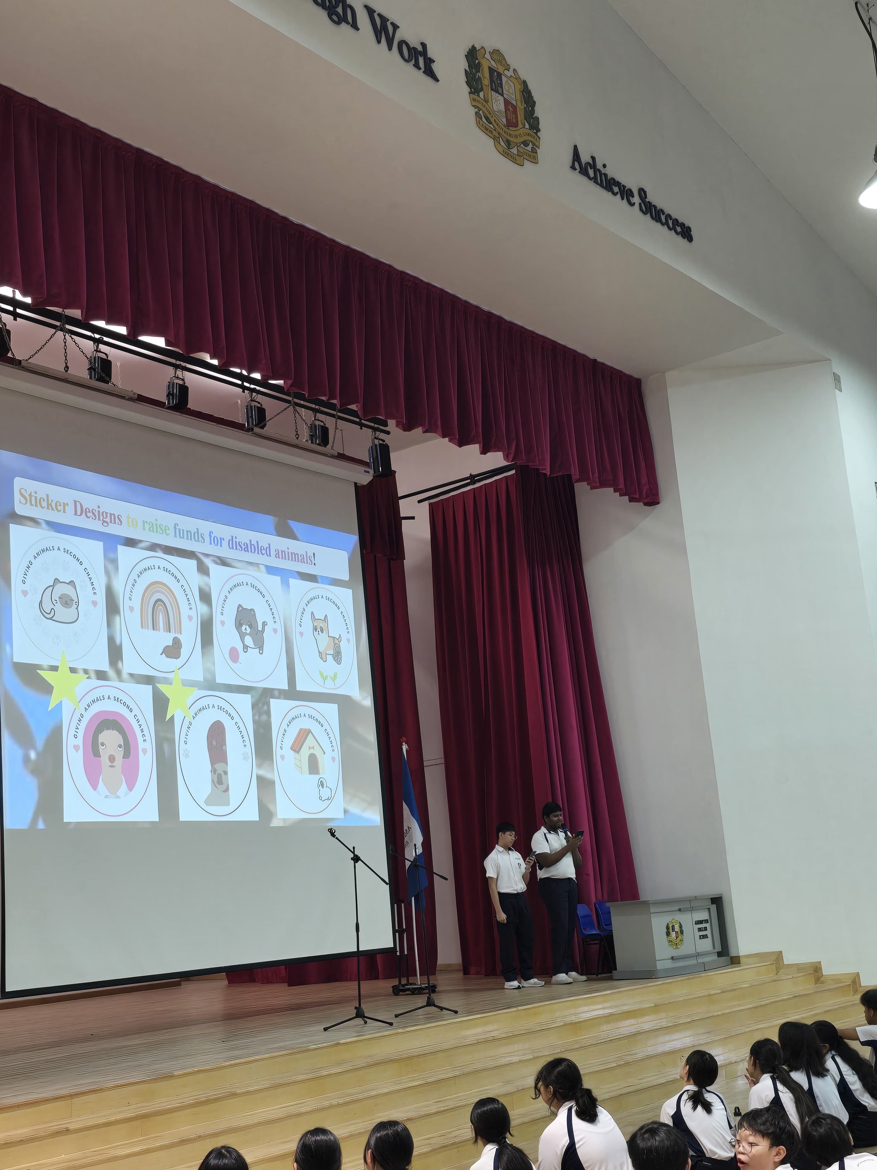 Two students present on stage showing a slideshow of sticker designs to raise funds for disabled animals.