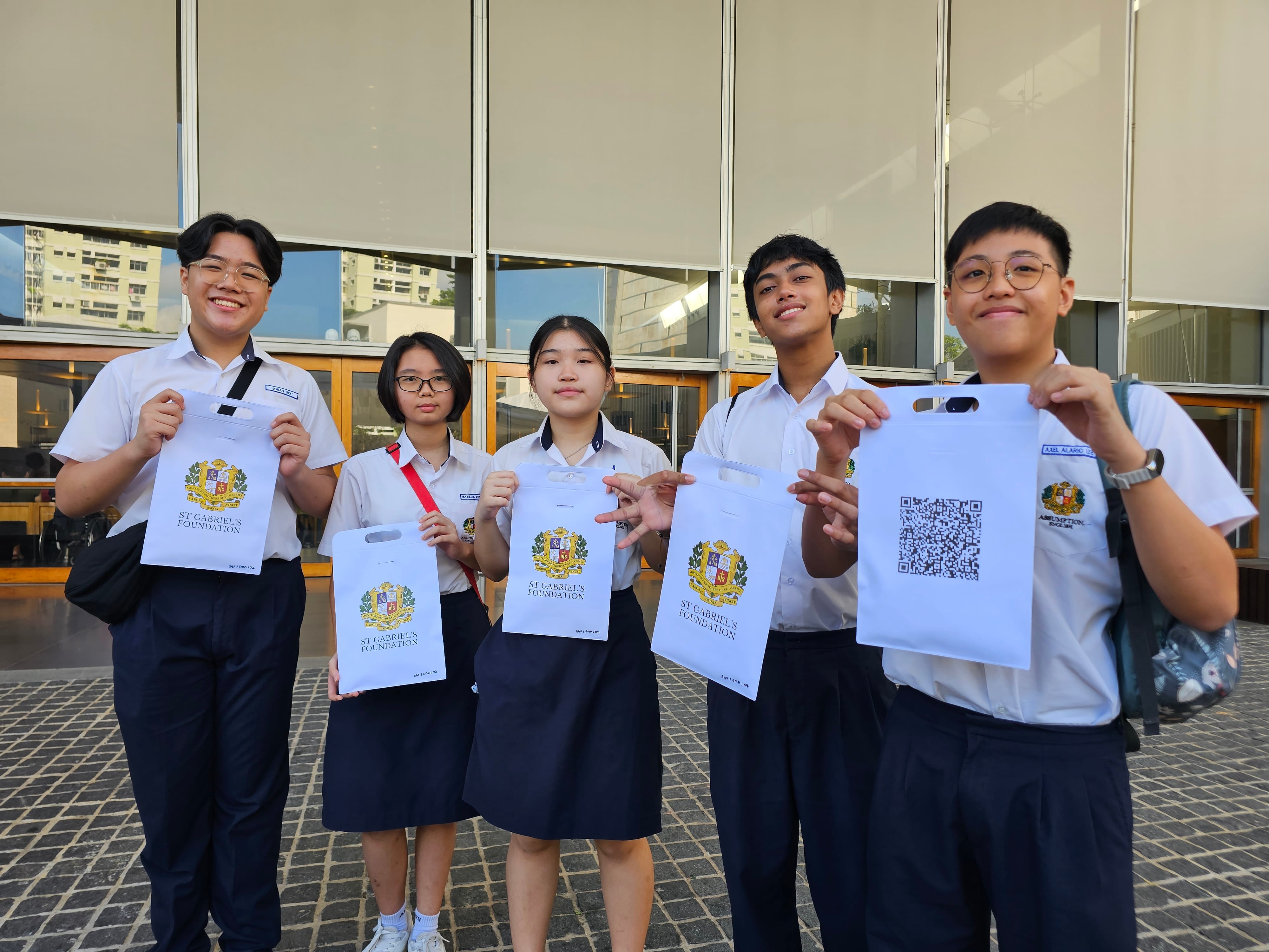Five students in school uniforms smile while holding white St Gabriel's Foundation bags outdoors.