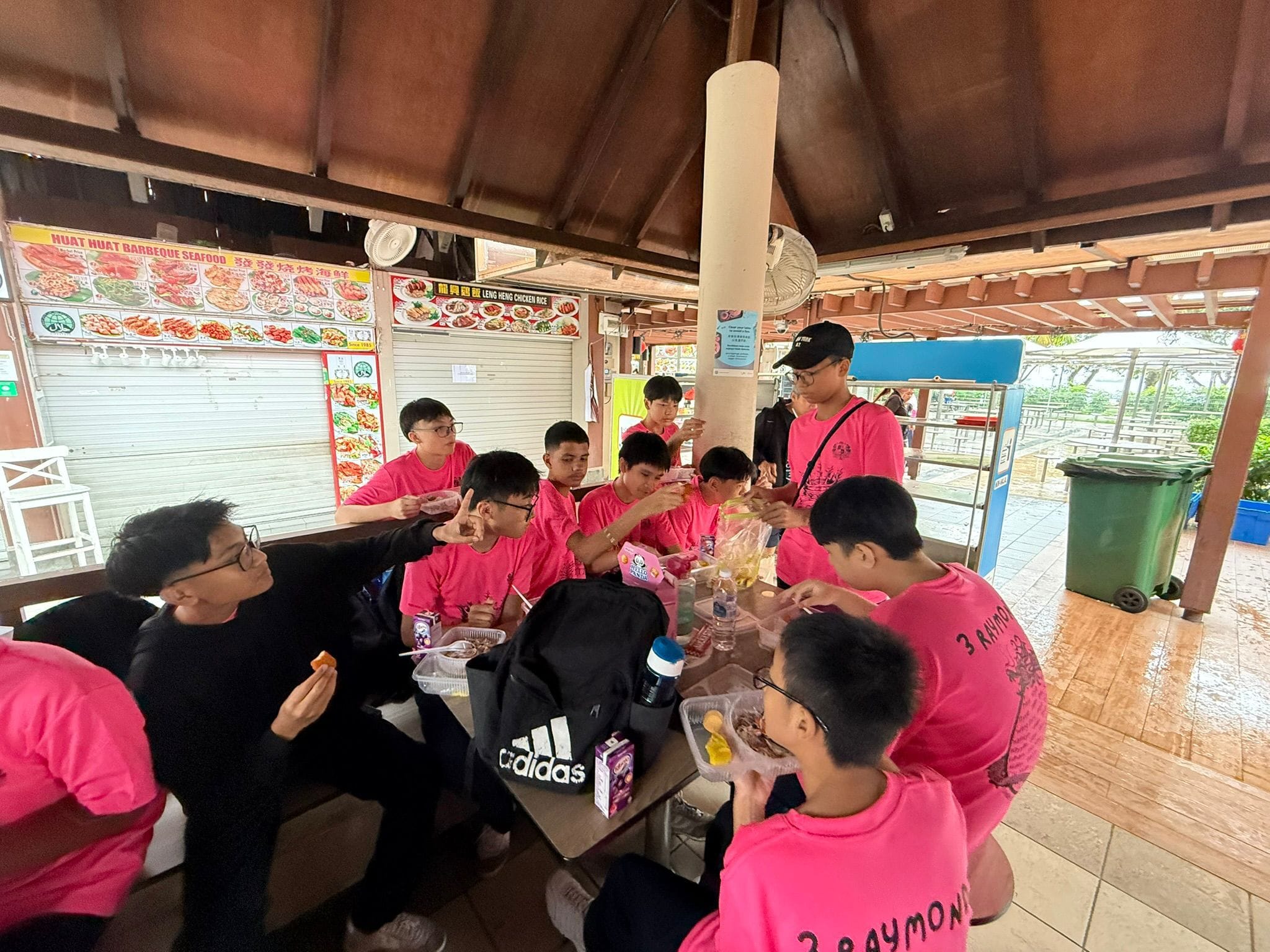 A group of young people in pink shirts eating at a table under a covered outdoor food court.