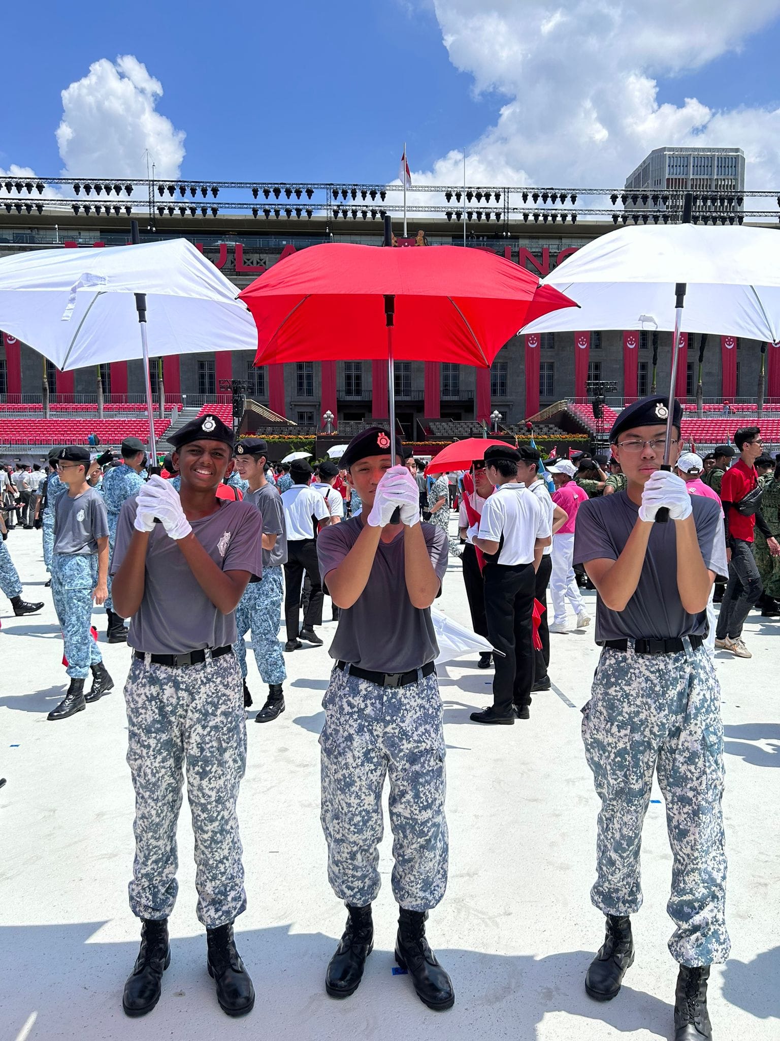Three uniformed cadets hold red and white umbrellas at an outdoor parade ground with a decorated stadium behind them.
