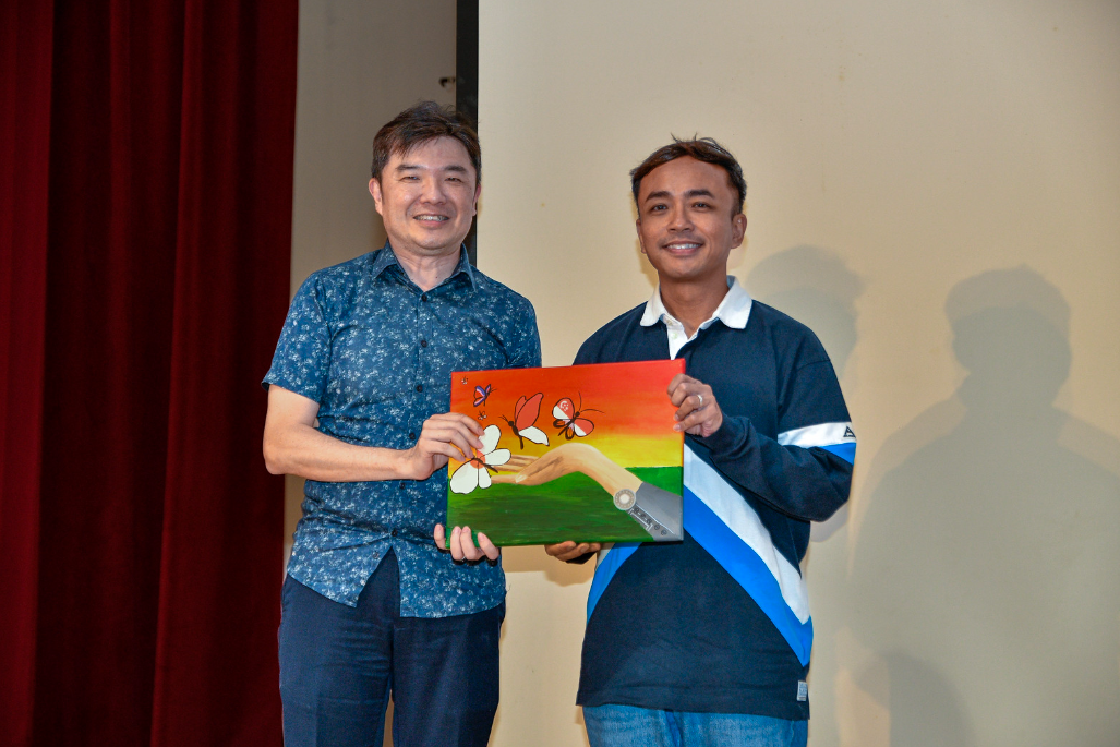Two smiling men hold a colorful painting featuring butterflies and hands on a rainbow background.