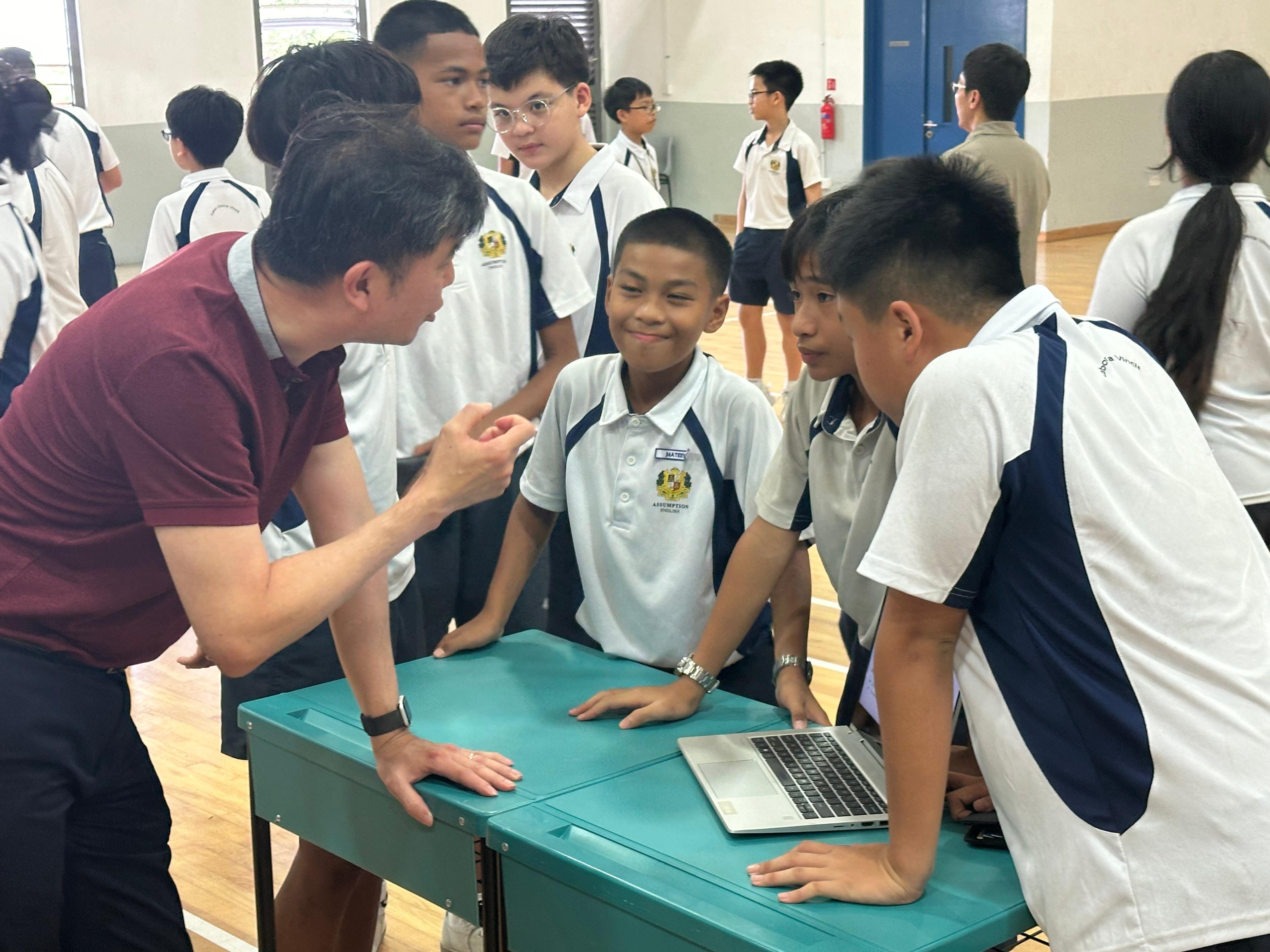 An adult in a red shirt talks to school students in uniforms gathered around a teal table with a laptop.