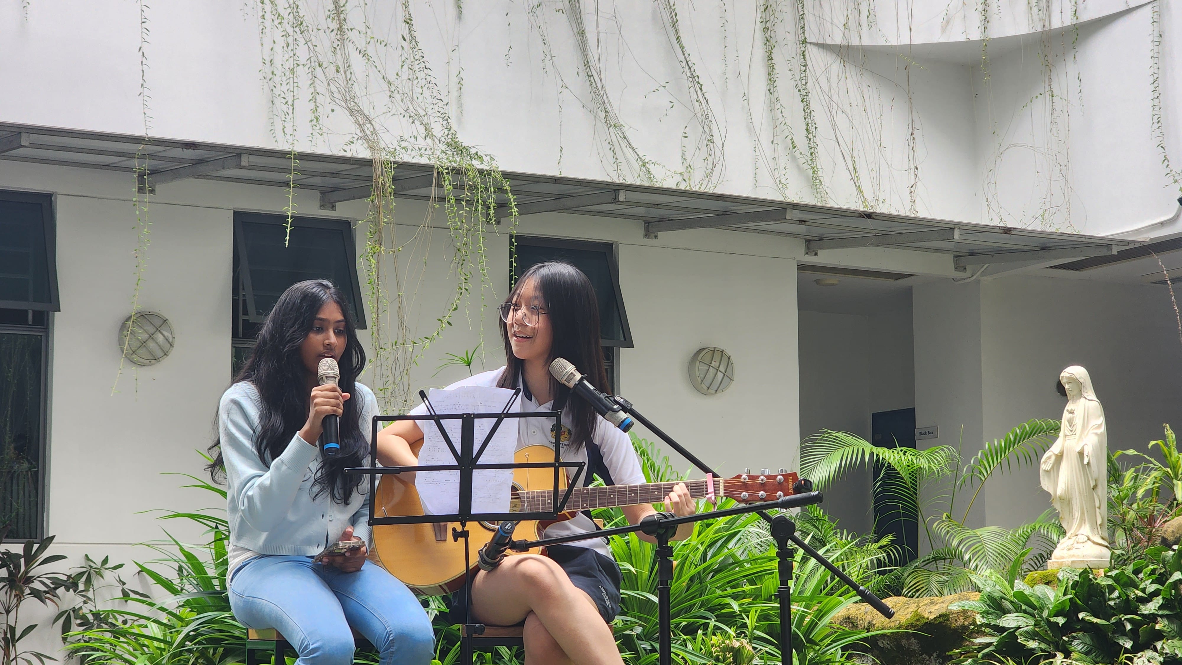 Two young women perform outdoors, one singing into a microphone and one playing acoustic guitar with sheet music on a stand.