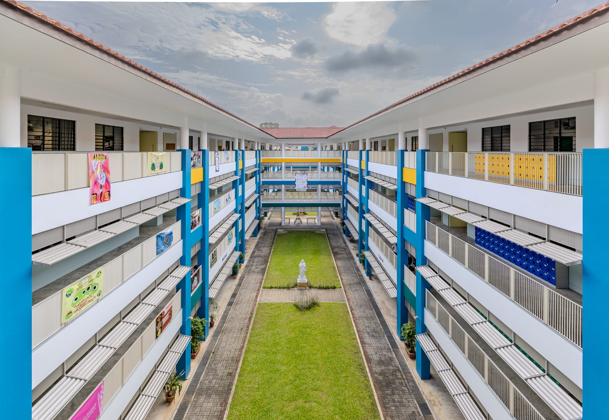 Multi-storey school courtyard with blue columns, green lawn, a white statue at center, and colorful posters on corridors.