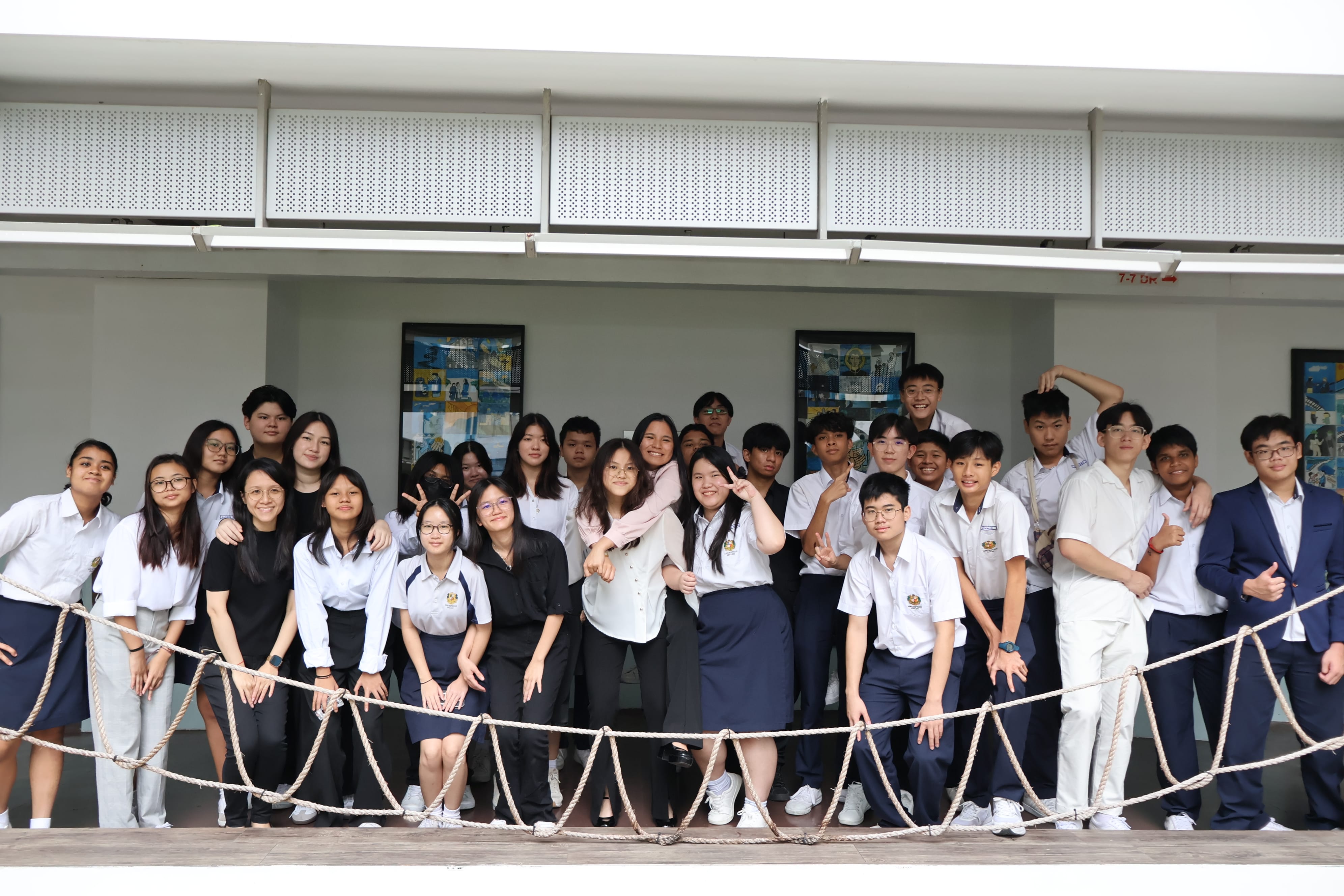 Group of students in school uniforms posing and smiling behind a rope railing in a corridor.