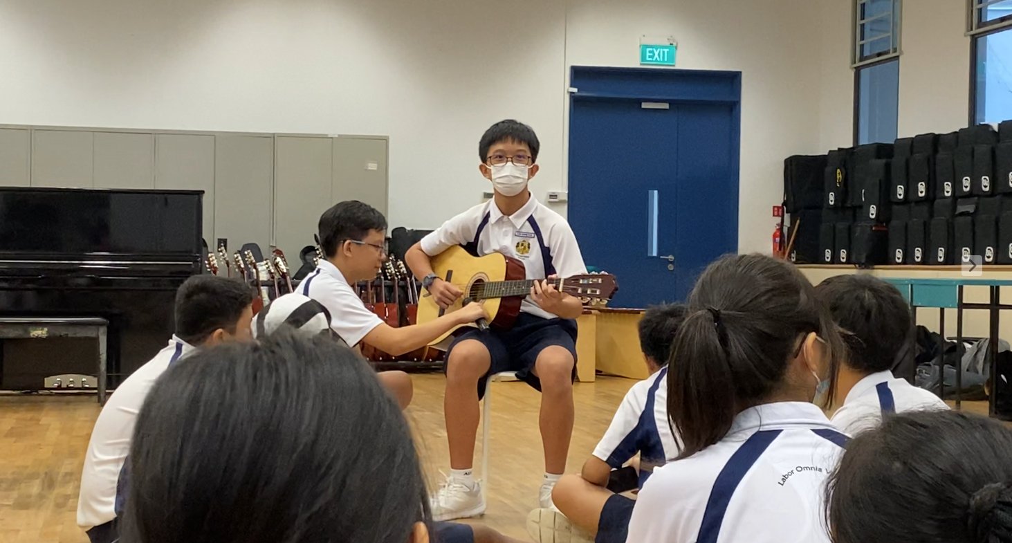 A masked student plays acoustic guitar while seated, surrounded by classmates in a school music room.
