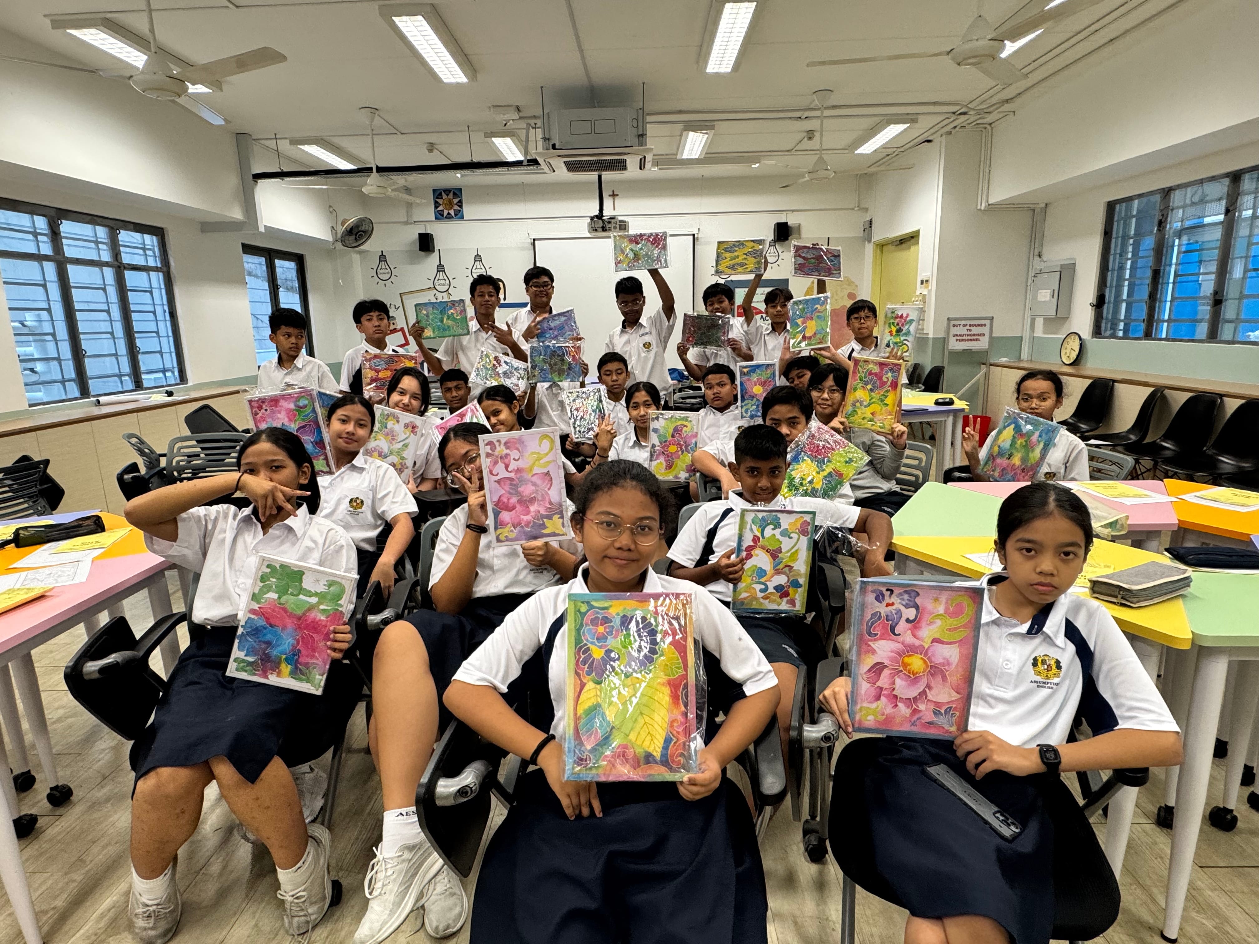 A group of school students in white uniforms hold up colorful floral artworks in a classroom.