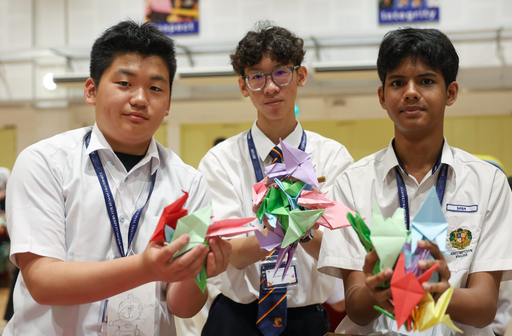 Three teenage boys in white school uniforms hold colorful paper origami cranes while smiling at the camera.