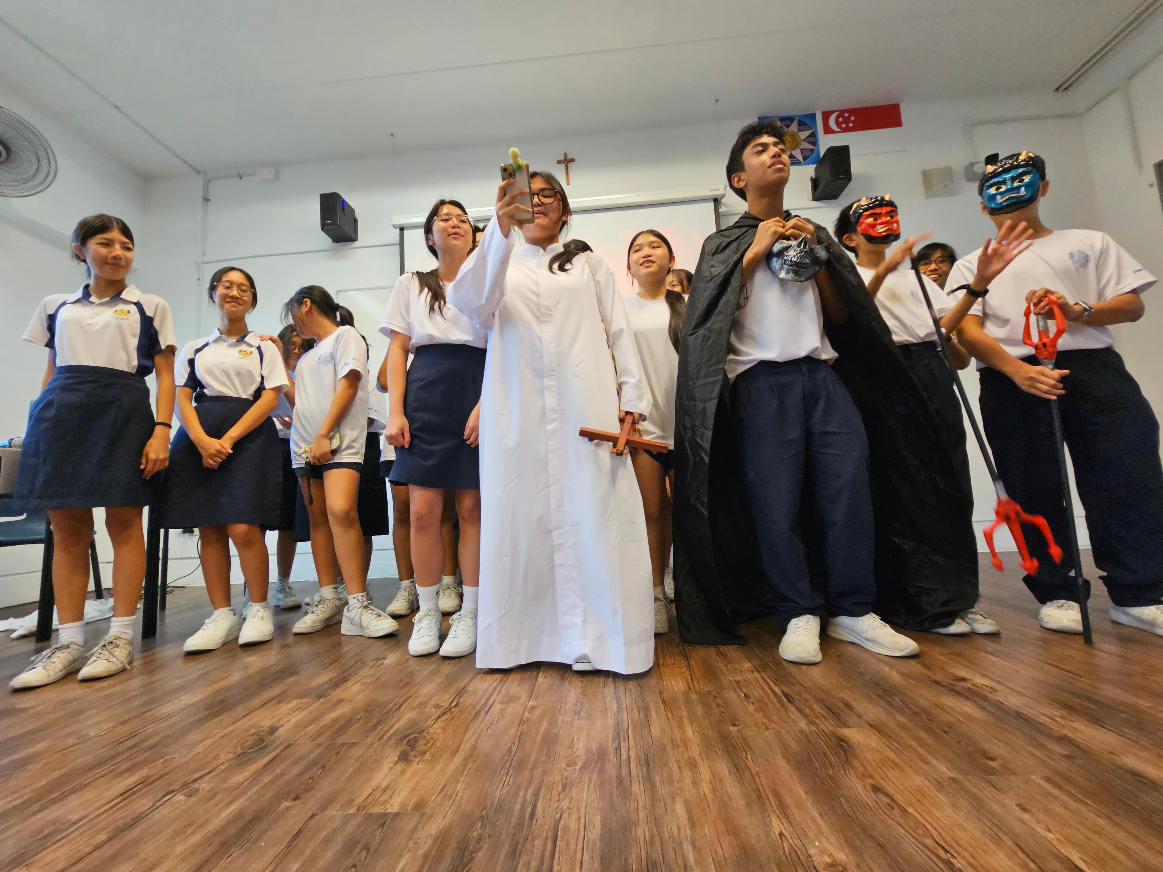 Students in uniforms perform a skit; one wears a white robe with a cross, another wears a black cape with a mask.
