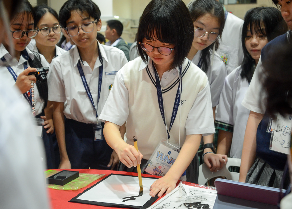 A student in uniform practices calligraphy with a brush while peers in lanyards watch closely.