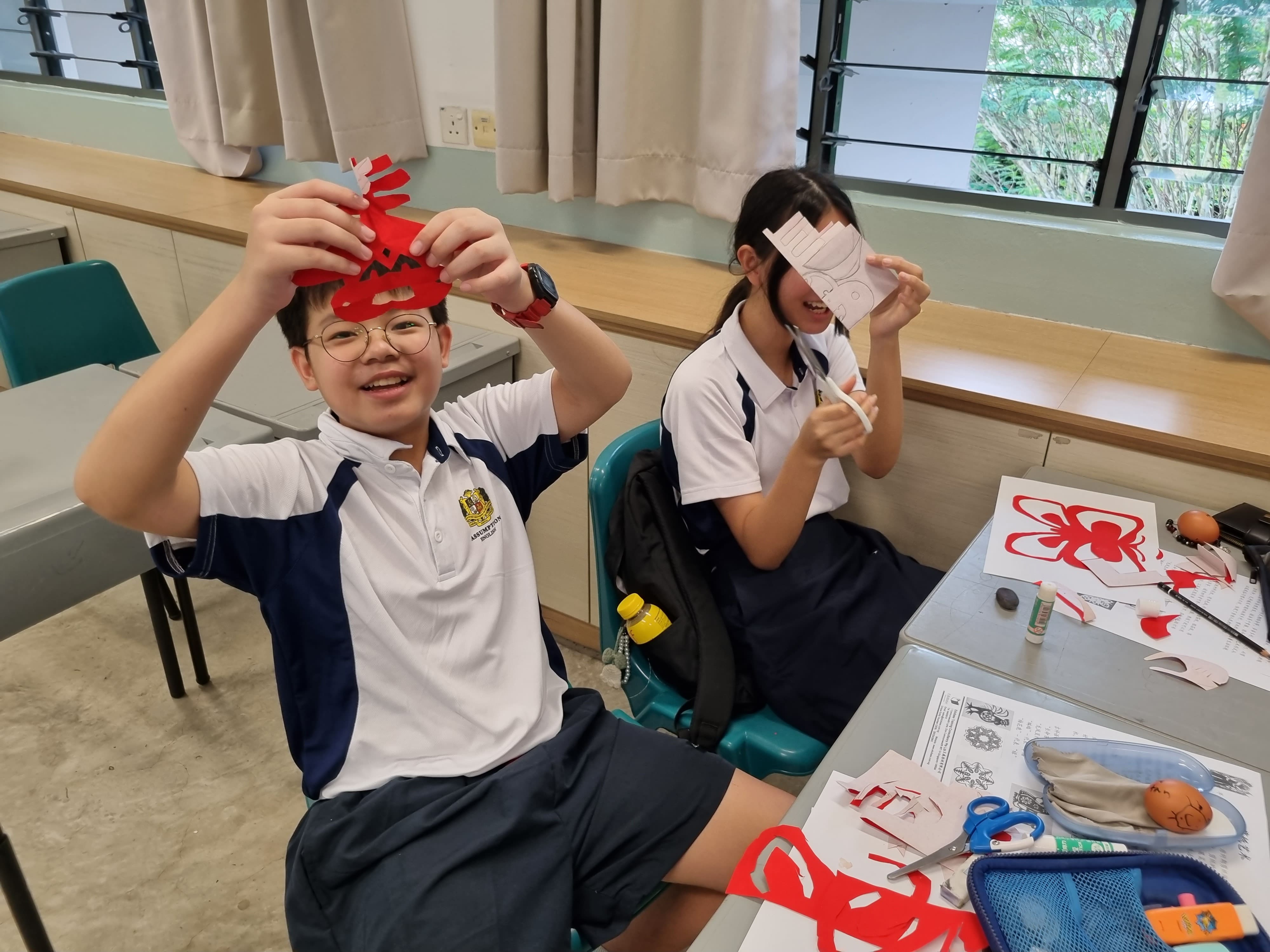 Two students in school uniforms hold up red paper-cut crafts and smile at a desk with scissors and craft supplies.
