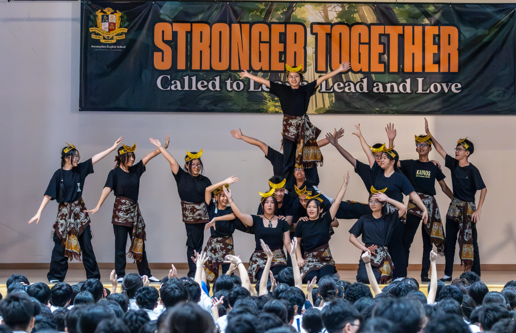 Students in black shirts and traditional headpieces perform on stage under a "Stronger Together" banner before a large audience.