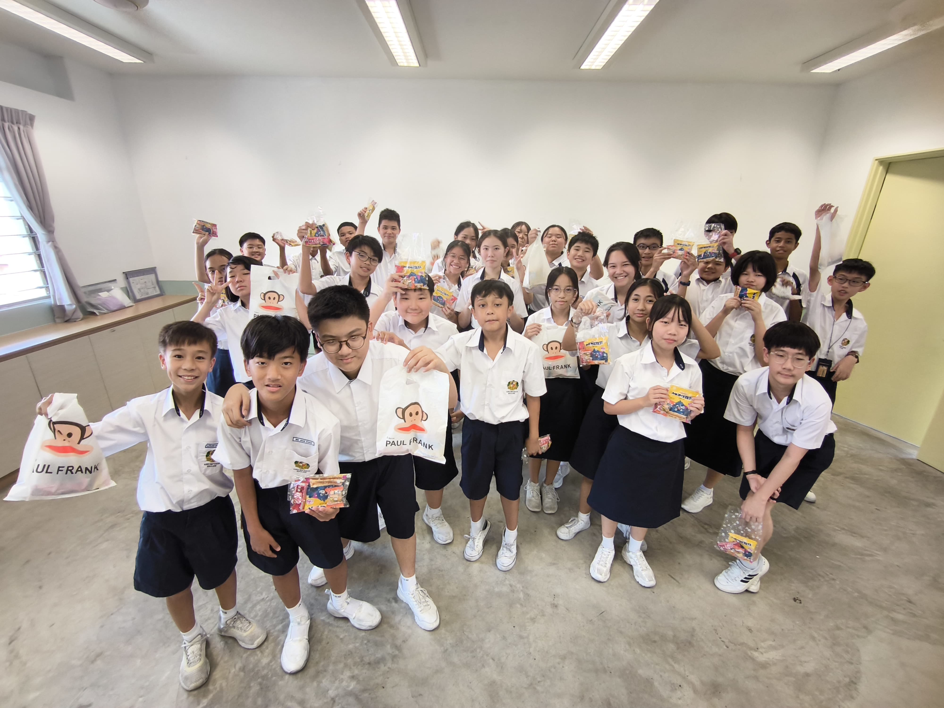 A large group of school students in white uniforms holding up snack packets and Paul Frank bags indoors.