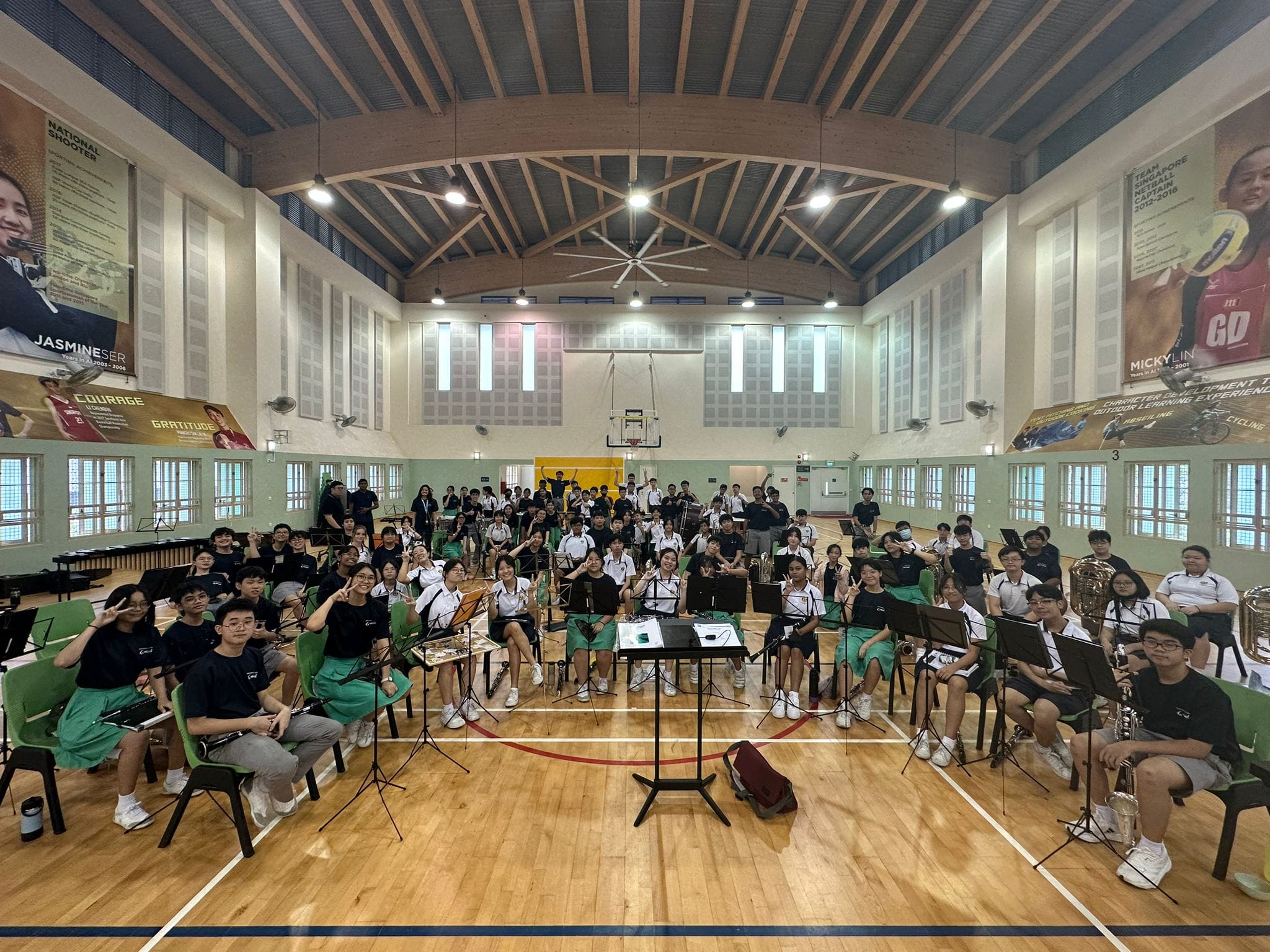 Large student orchestra seated with instruments and music stands in a school gymnasium with wooden ceiling.