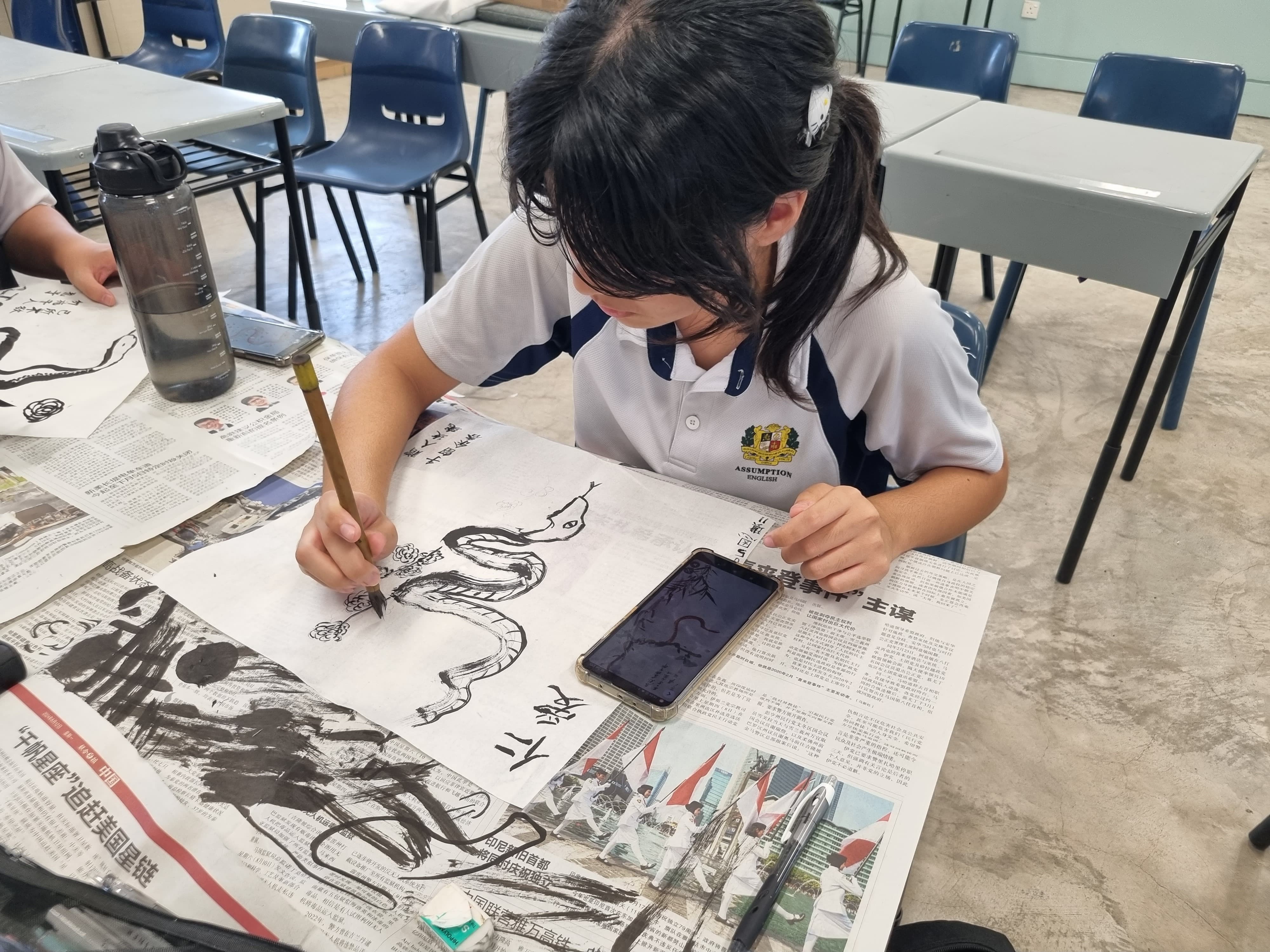 A student in a school uniform paints a black ink snake on paper spread over a newspaper-covered desk.