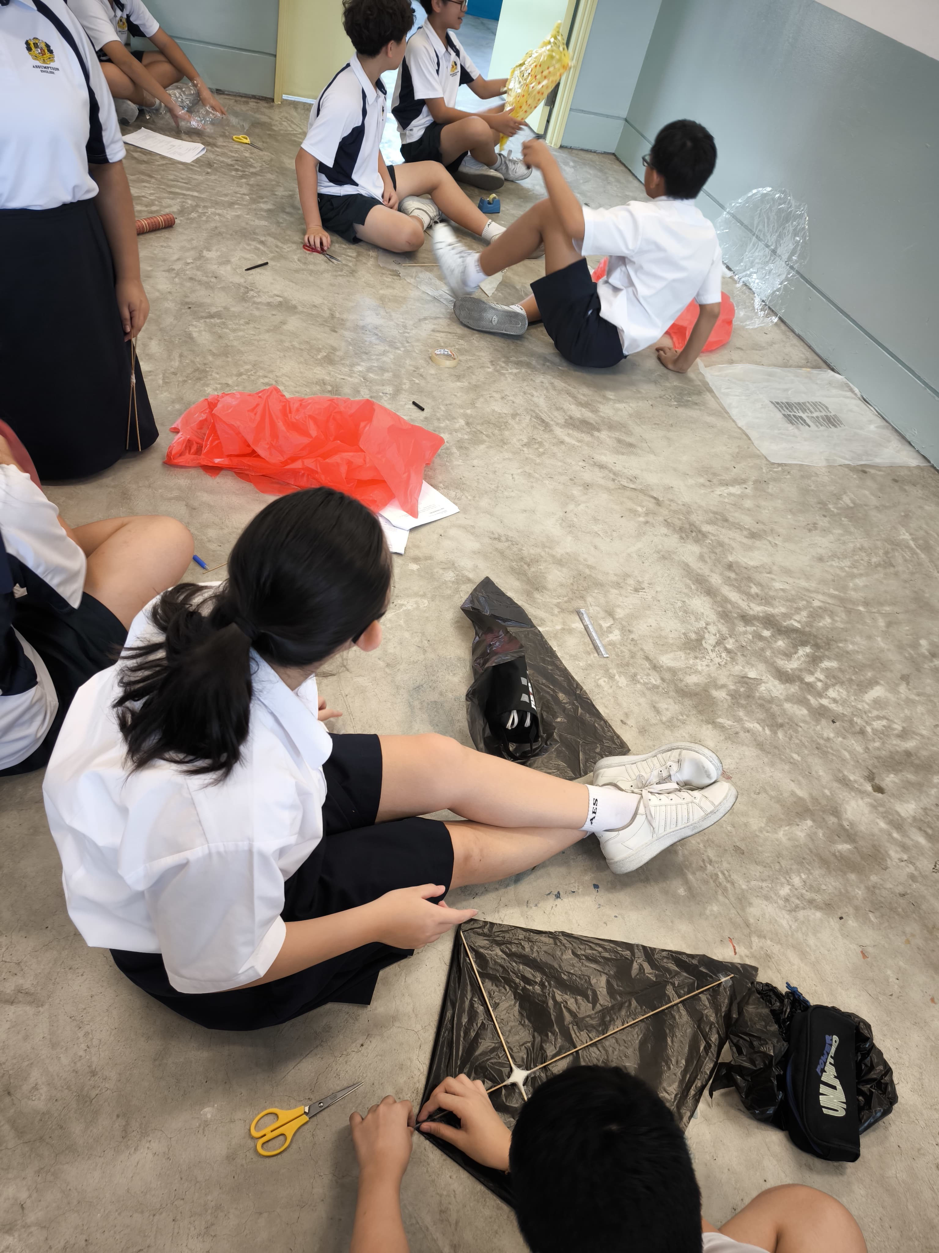 Students in white uniforms sit on a floor building kites from plastic bags and sticks using scissors.